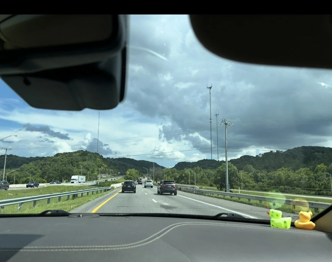View through a car windshield of a highway leading toward distant mountains under a cloudy sky.