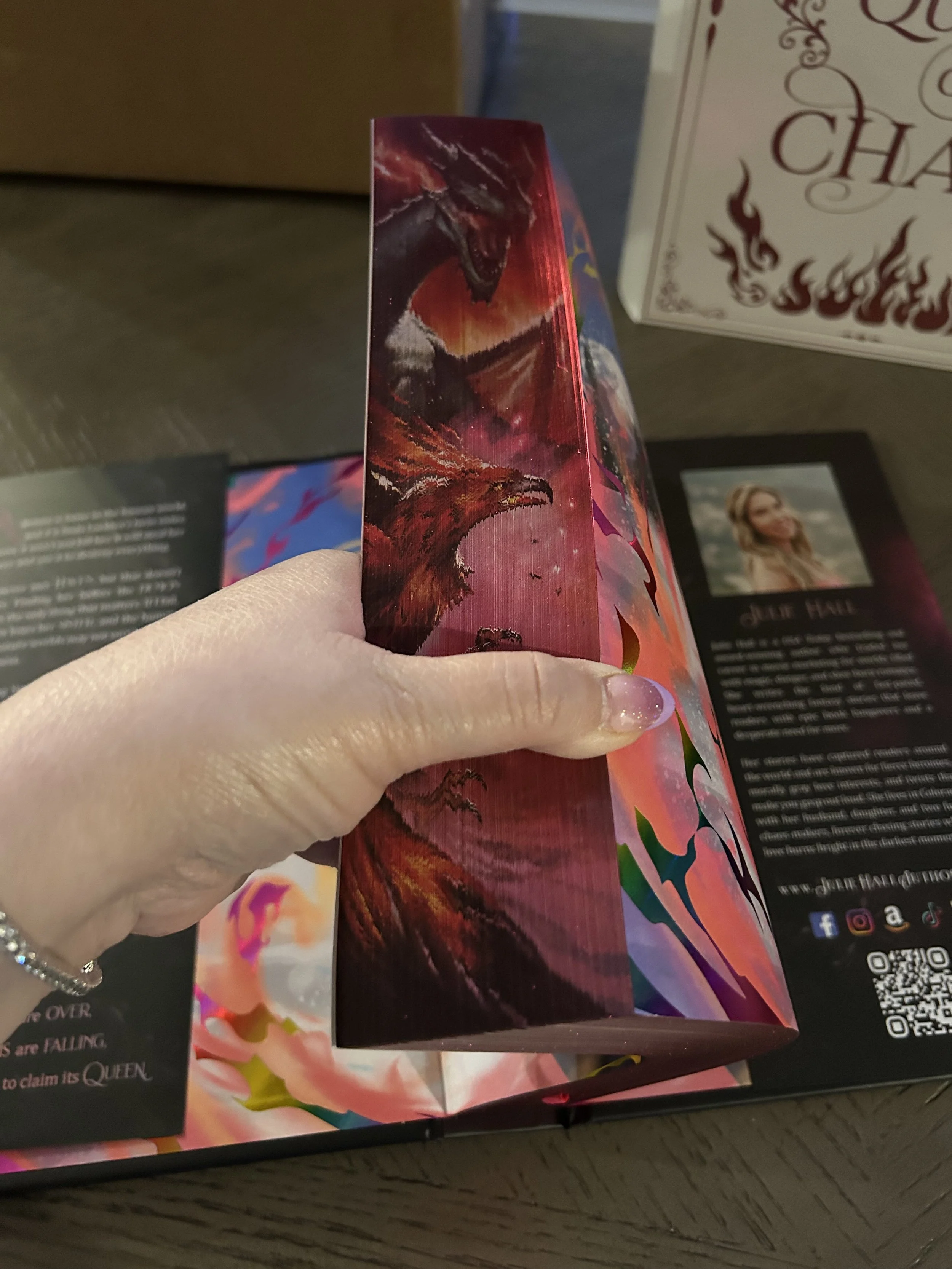 Decorated page edge of Queen of Chaos featuring several dragons against a shimmering red and pink fantasy background.