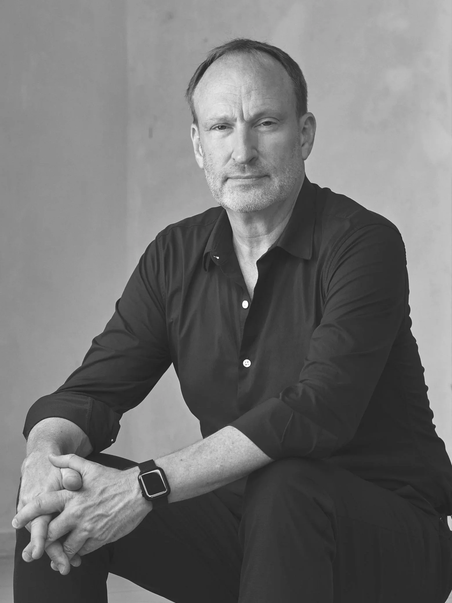 Black and white photo of a middle-aged man with short hair, some facial hair, wearing a dark button-up shirt, sitting with his arms crossed, looking directly at the camera.