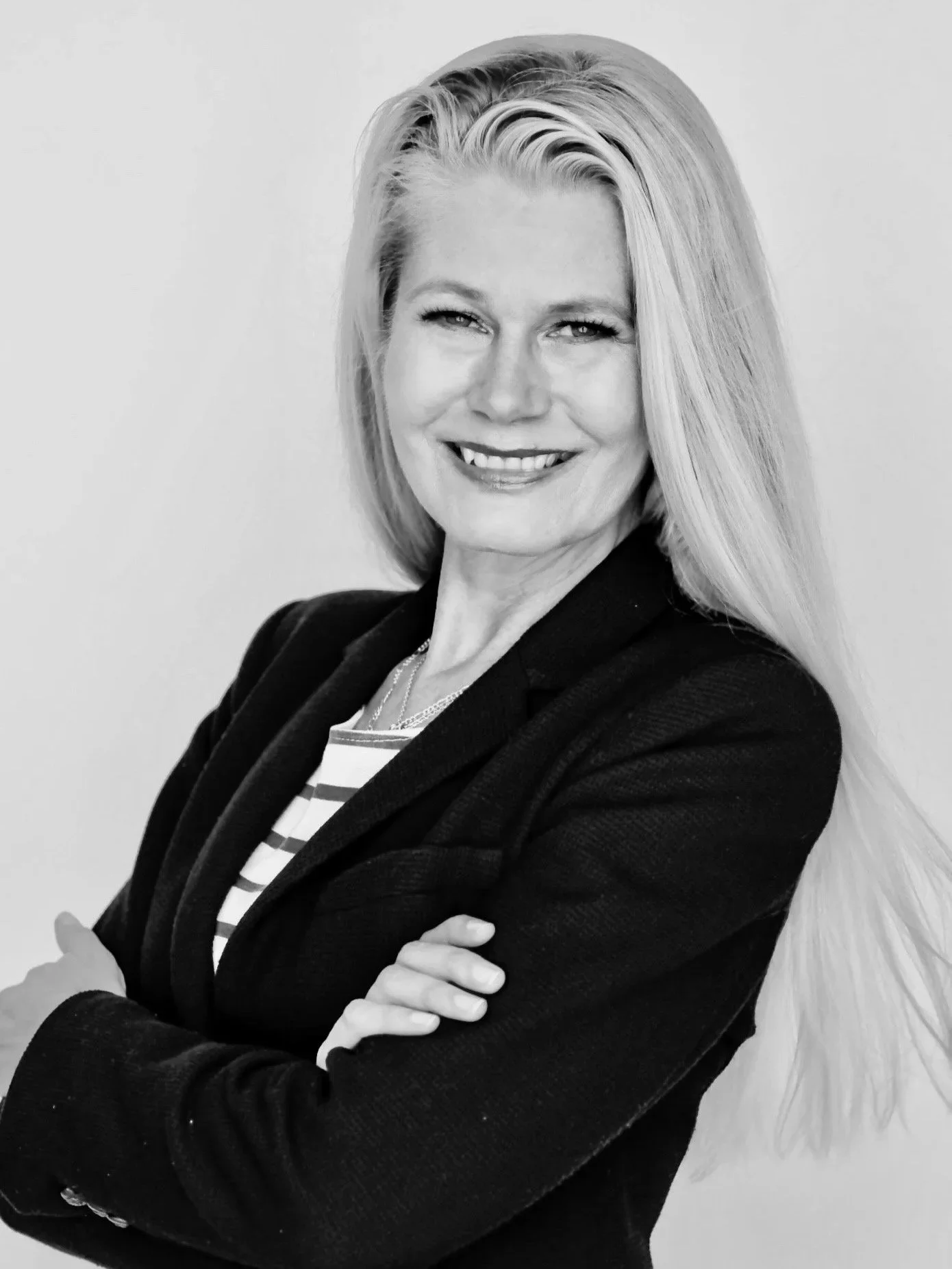 Black and white portrait of a smiling woman with long light-colored hair, crossed arms, wearing a striped shirt and a dark blazer, standing against a plain background.