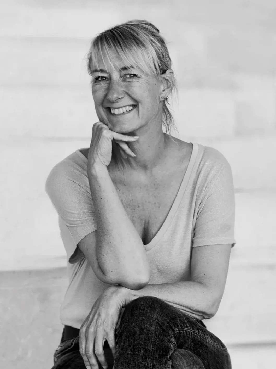 A smiling woman with short, light-colored hair, sitting with her chin resting on her hand, wearing a light-colored V-neck shirt and dark jeans, in black and white.