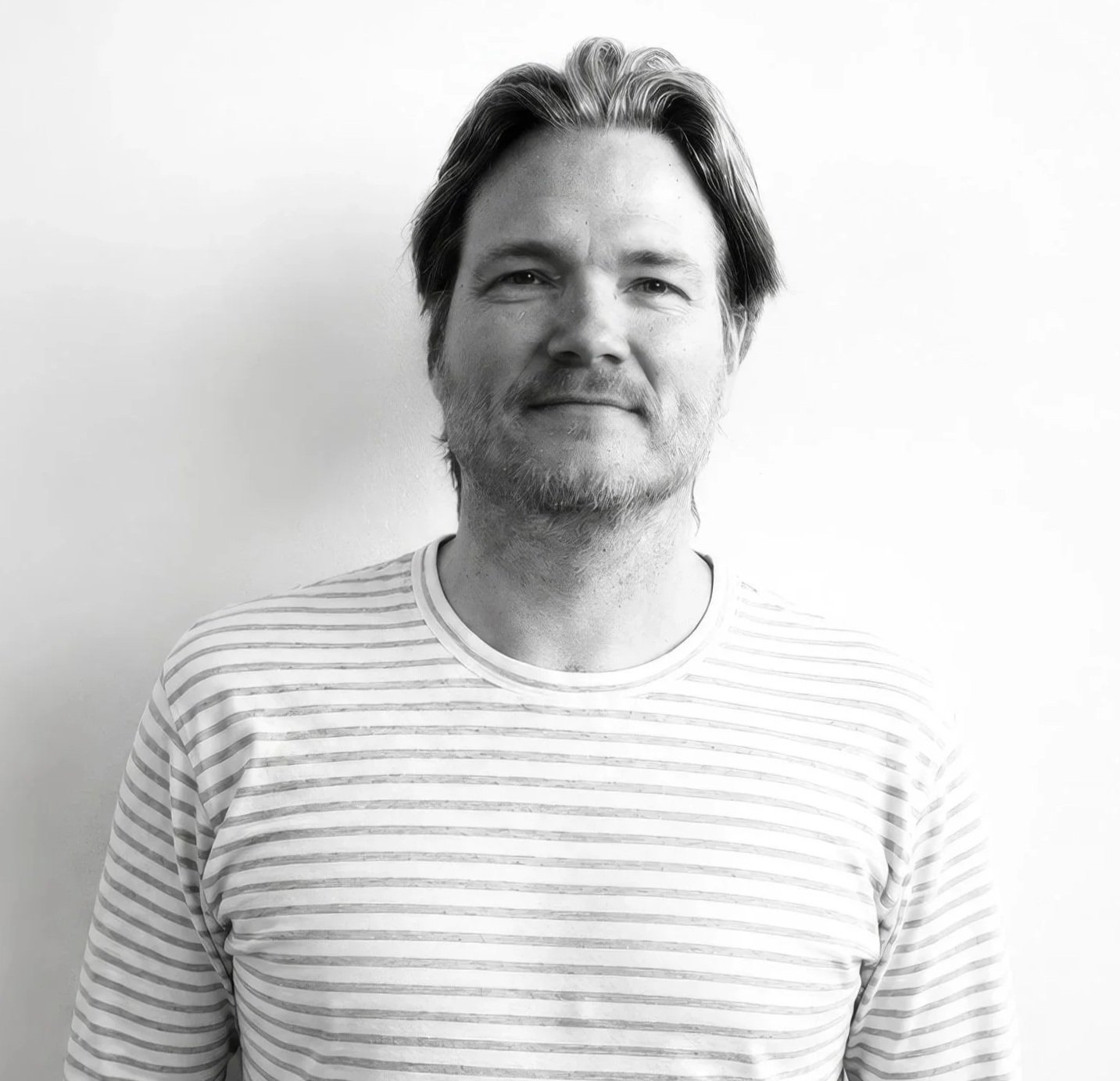 Black and white portrait of a man with shoulder-length hair, slight beard, and mustache, wearing a striped shirt, standing against a plain white background.
