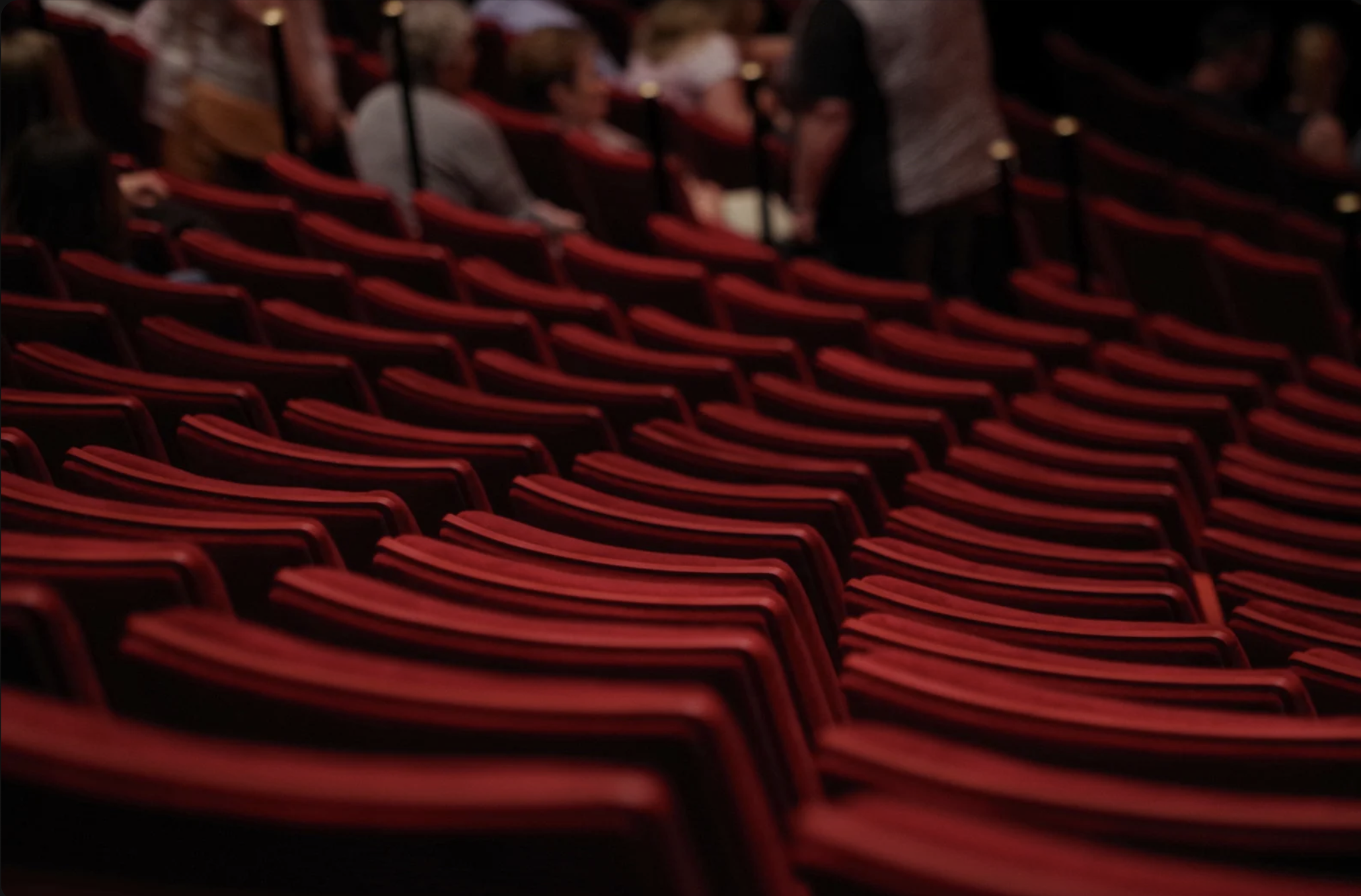 Empty red theater seats with a few people seated in the background and some people standing.