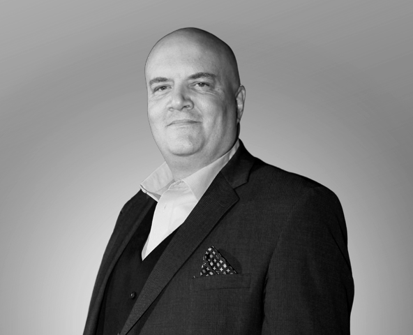 A black and white portrait of a bald man wearing a dark suit jacket, white shirt, and a patterned pocket square, looking confidently at the camera.