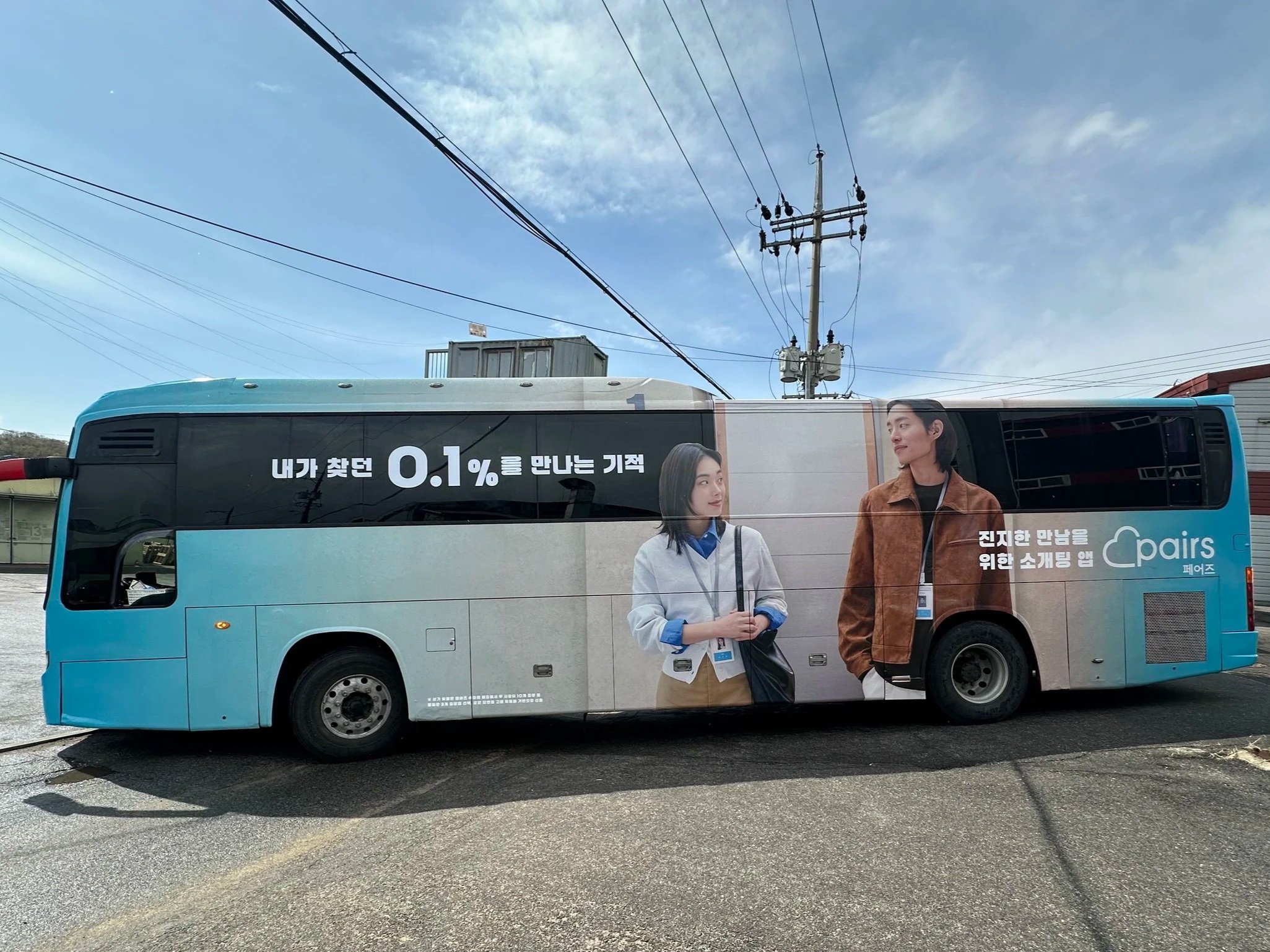 A light blue bus with advertisement featuring two people, a woman and a man, standing outdoors under a cloudy sky with power lines overhead.