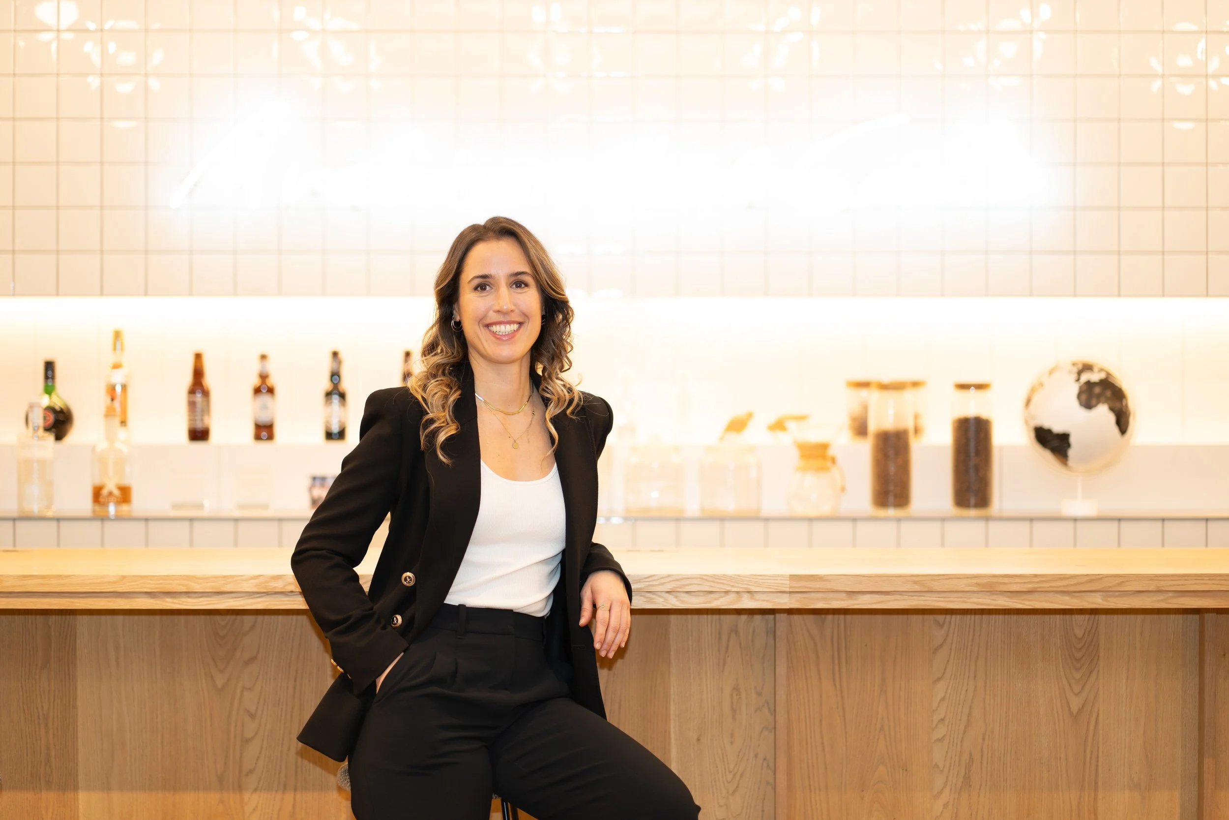 A woman with wavy brown hair smiling, dressed in a black blazer and white top, sitting at a wooden counter in a modern, minimalistic setting with bottles and glass jars behind her.
