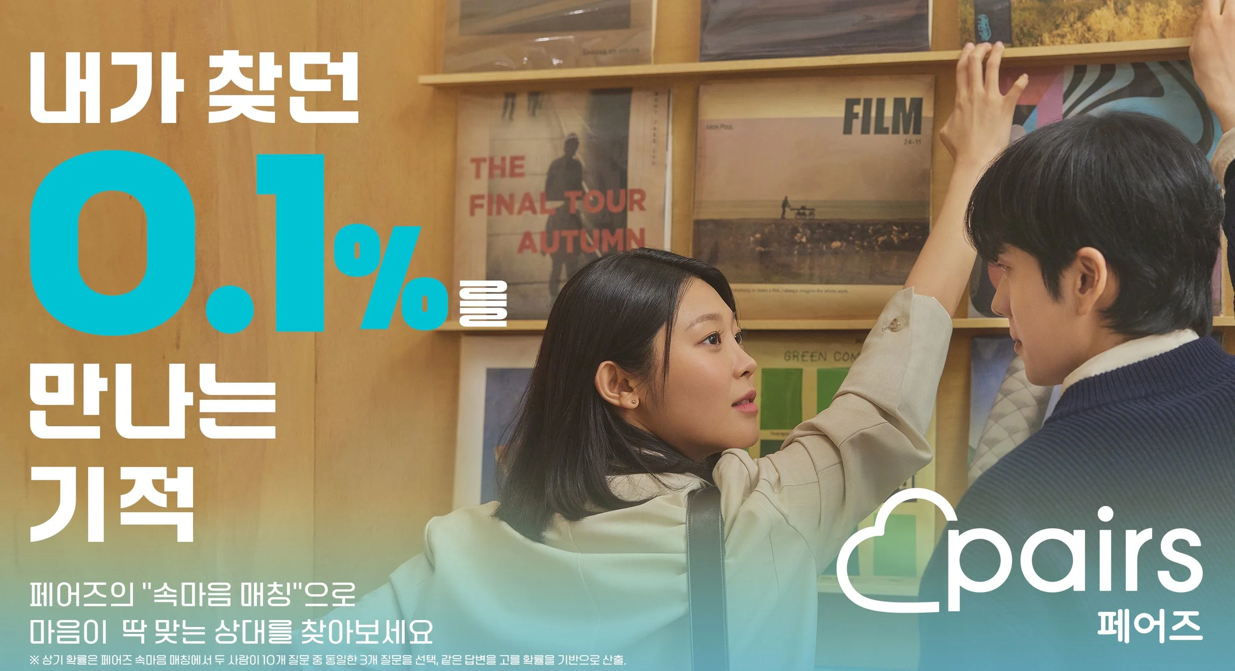Two people, a woman and a man, are in a bookstore looking at records on a shelf. The woman is reaching for a record while the man watches. There are various records on the shelf behind them.