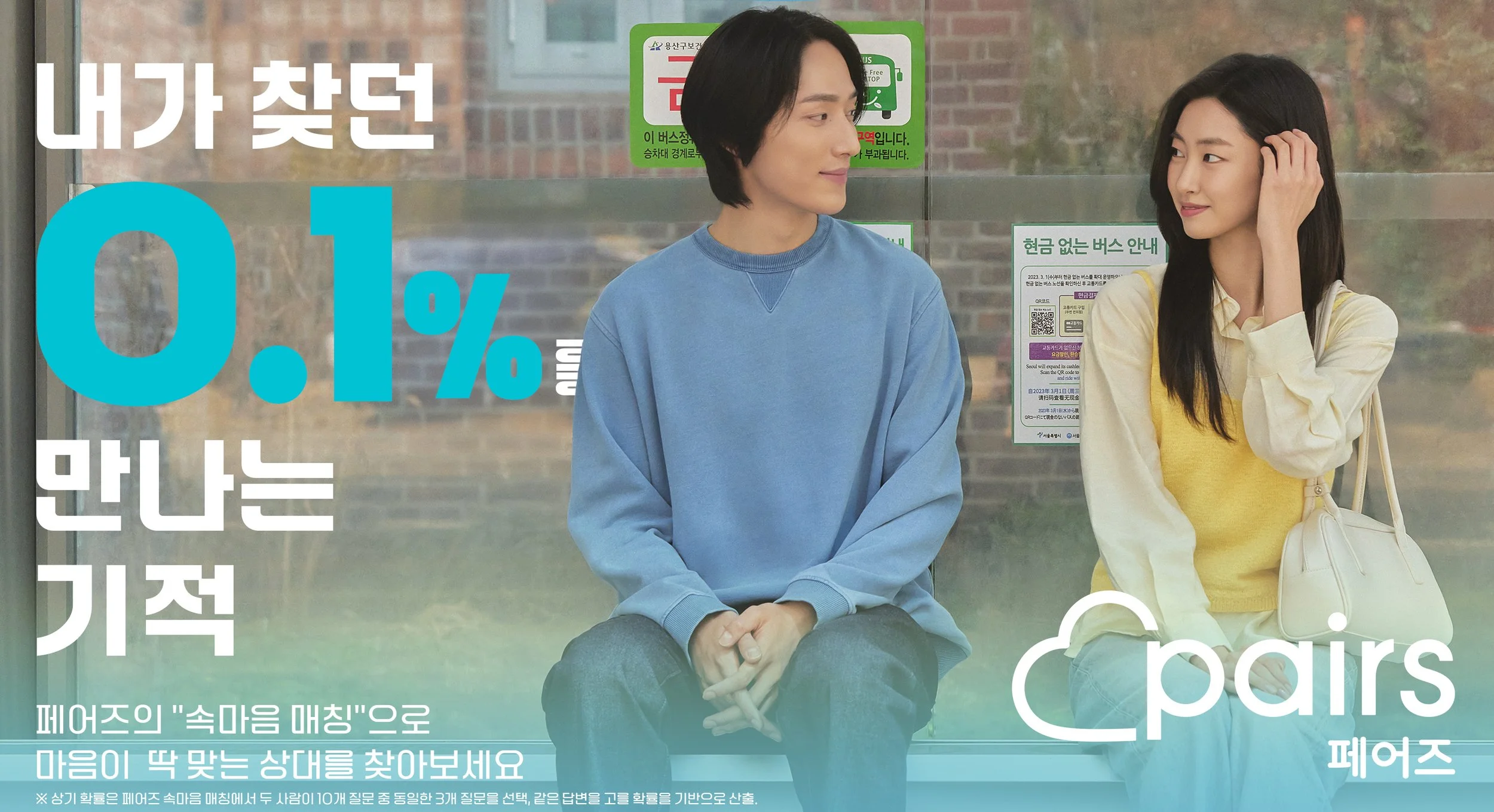 Two women sitting on a bench at a bus stop, engaging in conversation. One woman is wearing a blue sweatshirt, and the other is wearing a yellow vest with a cream-colored jacket. Behind them, there are signs and notices on the glass, including a green