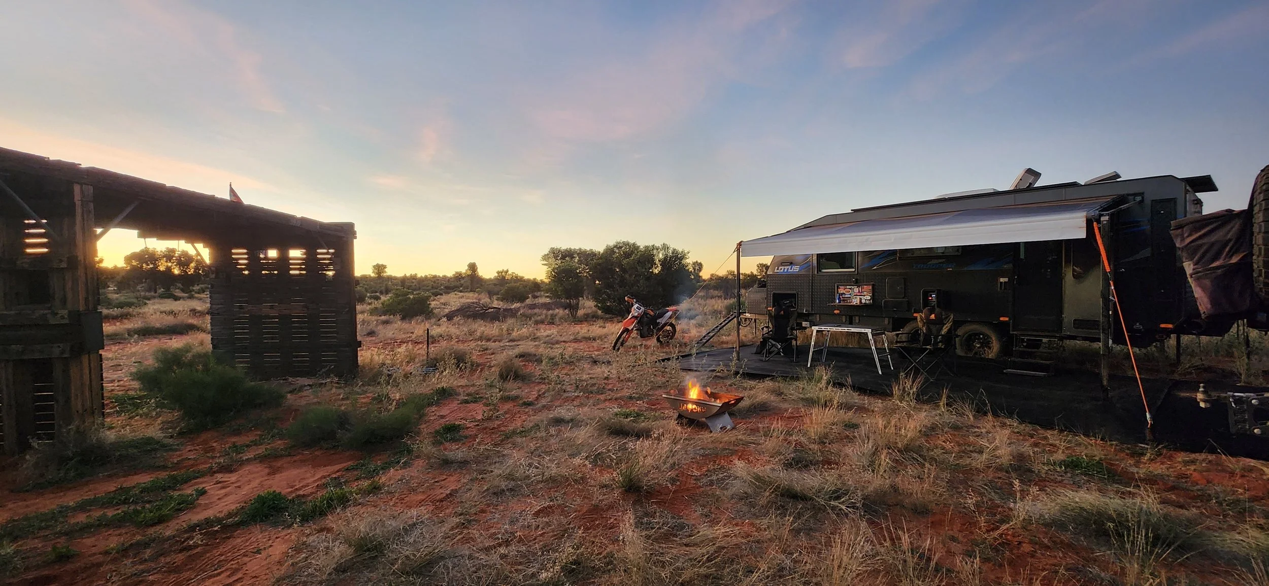 A camping scene at sunset with a trailer, a motorbike, a small fire pit, and camping chairs in a desert landscape with sparse vegetation and a wooden structure.