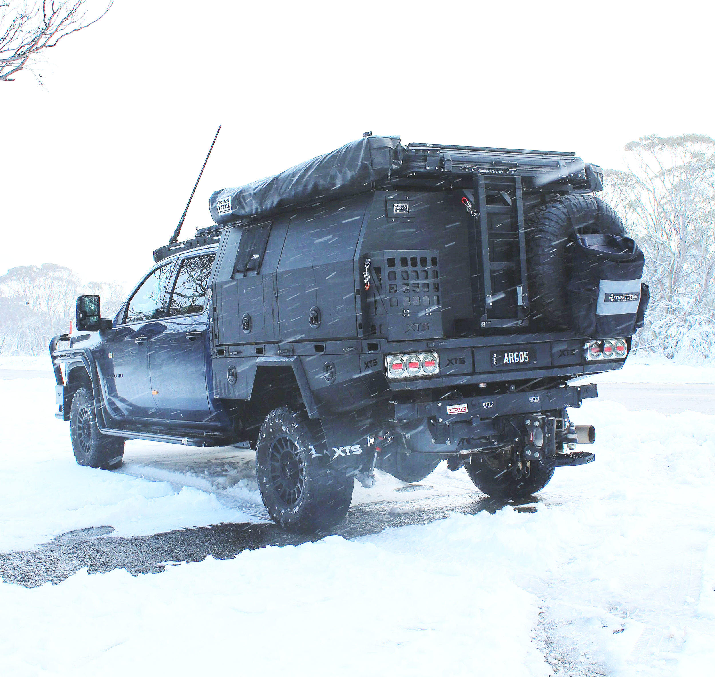 Black off-road vehicle with roof storage and large tires parked in snowy landscape with trees in the background.