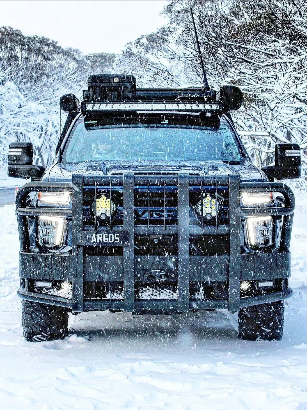 Black off-road vehicle with heavy-duty front bumper, grille guard, and mounted auxiliary lights, driving through snowy landscape with snow-covered trees in the background.