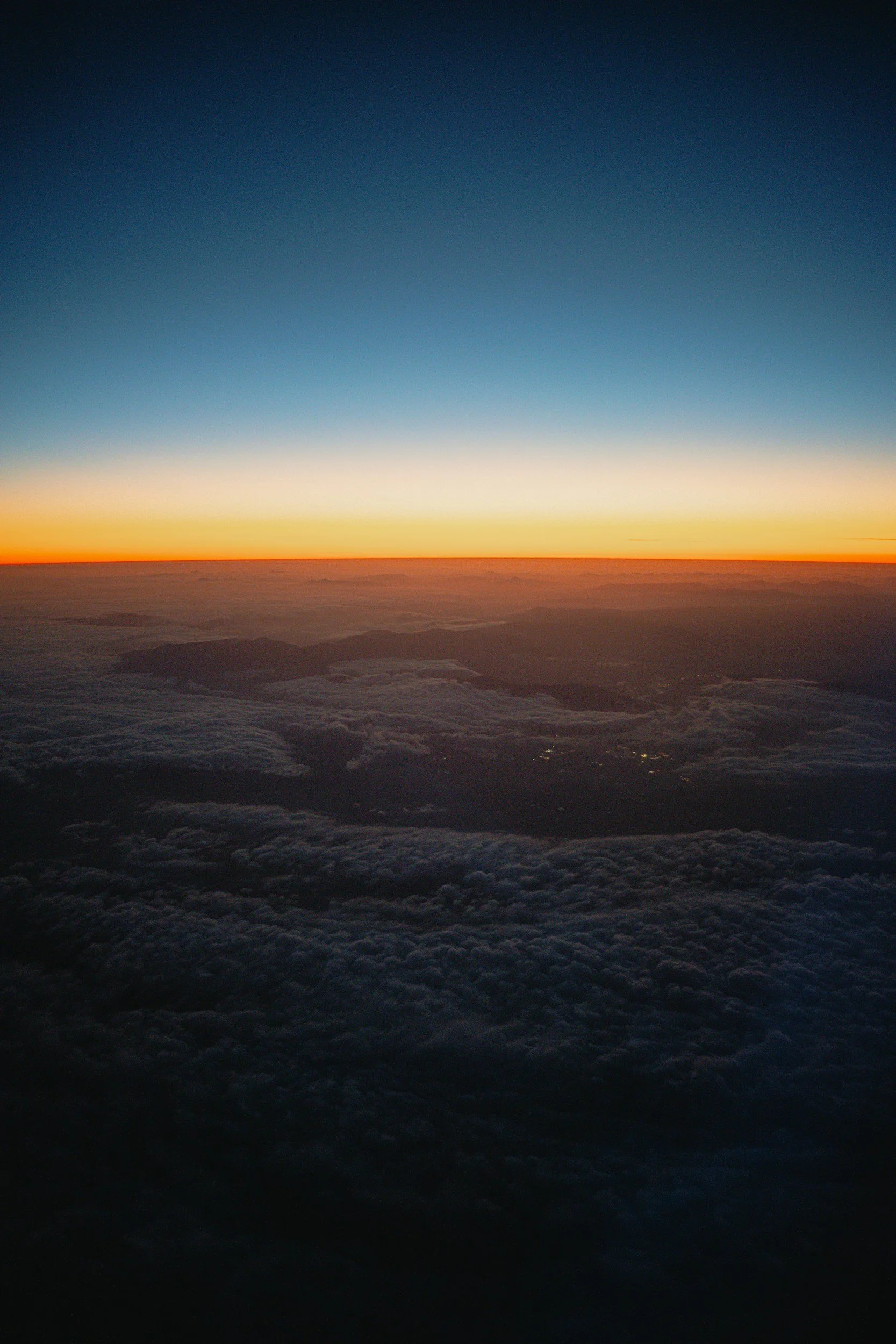 View of the sunset or sunrise over a mountain range, with a sky transitioning from deep blue to orange, and clouds below.