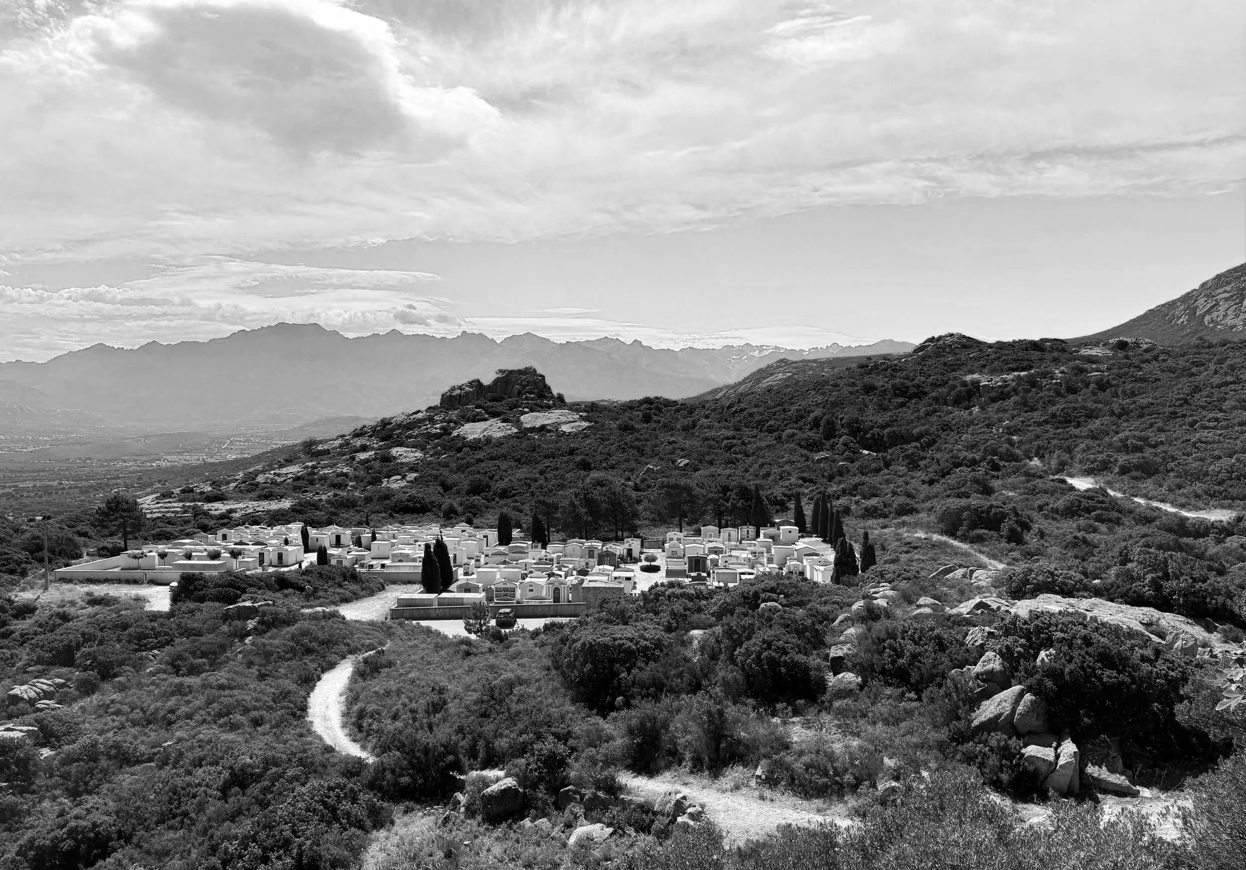 Un paysage de montagne avec des collines verdoyantes, un petit village avec des maisons blanches, et des montagnes au loin, en noir et blanc.