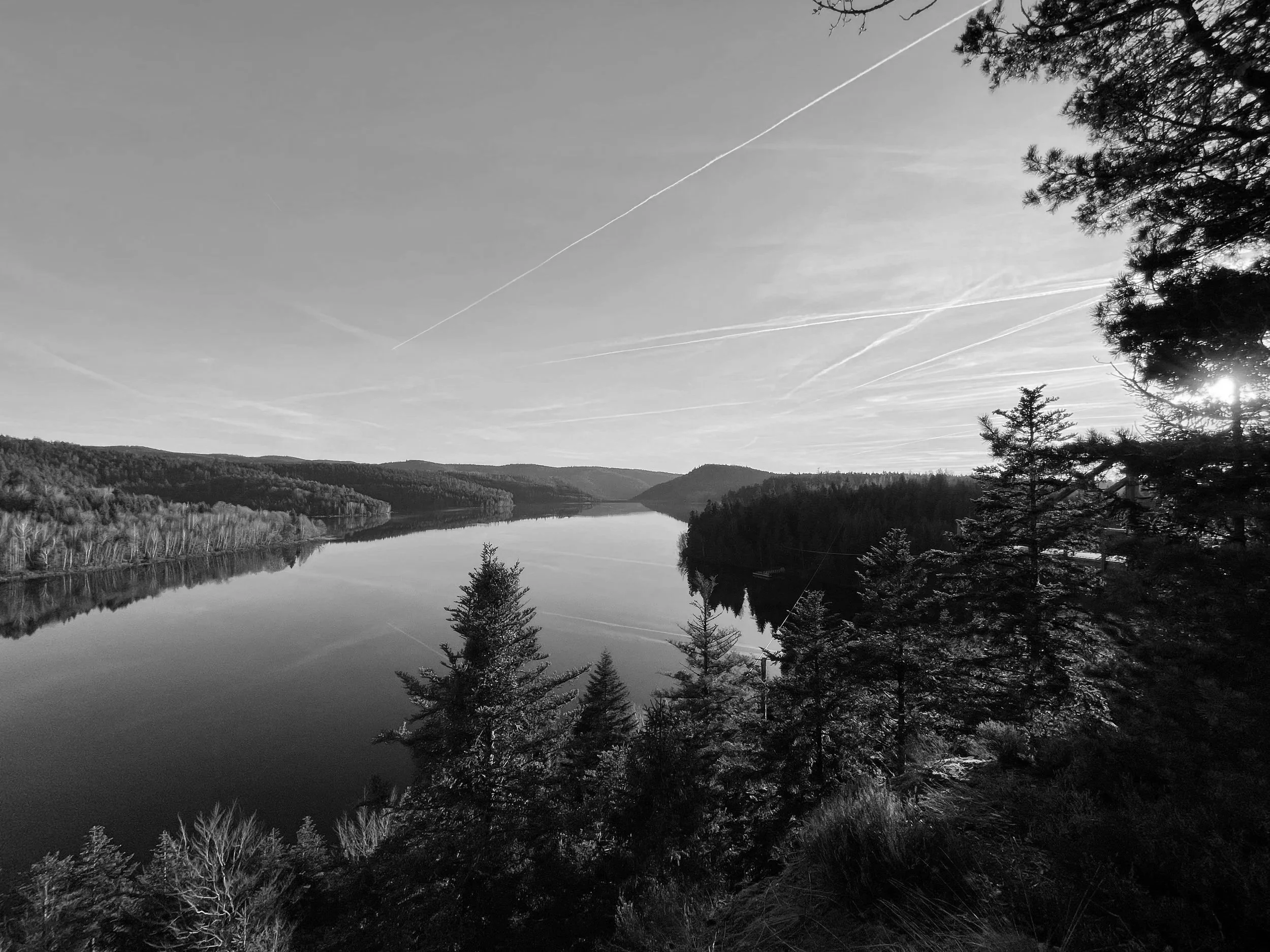 Paysage en noir et blanc avec un lac entouré de forêts et de collines, avec un ciel clair marqué par plusieurs traînées d'avions.