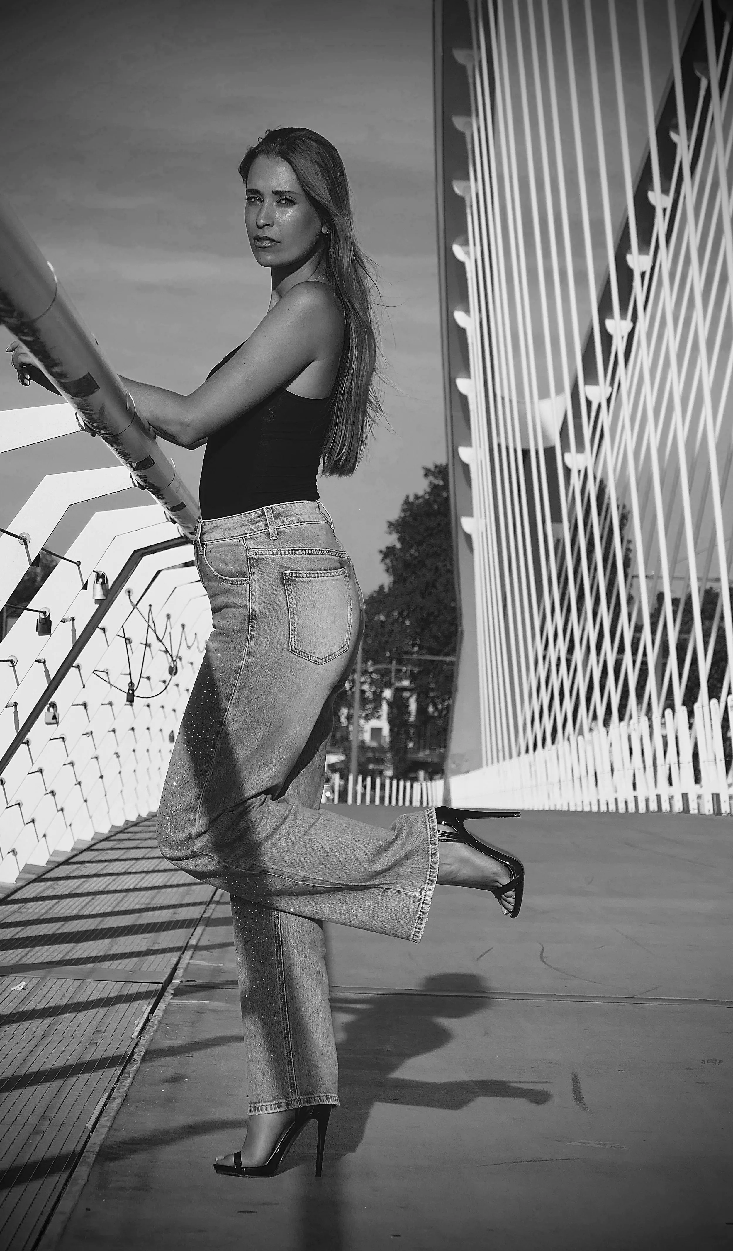 Une femme porte un haut noir, un jean délavé, et des talons hauts noirs, posant sur un pont avec une structure métallique blanche et un ciel en arrière-plan.