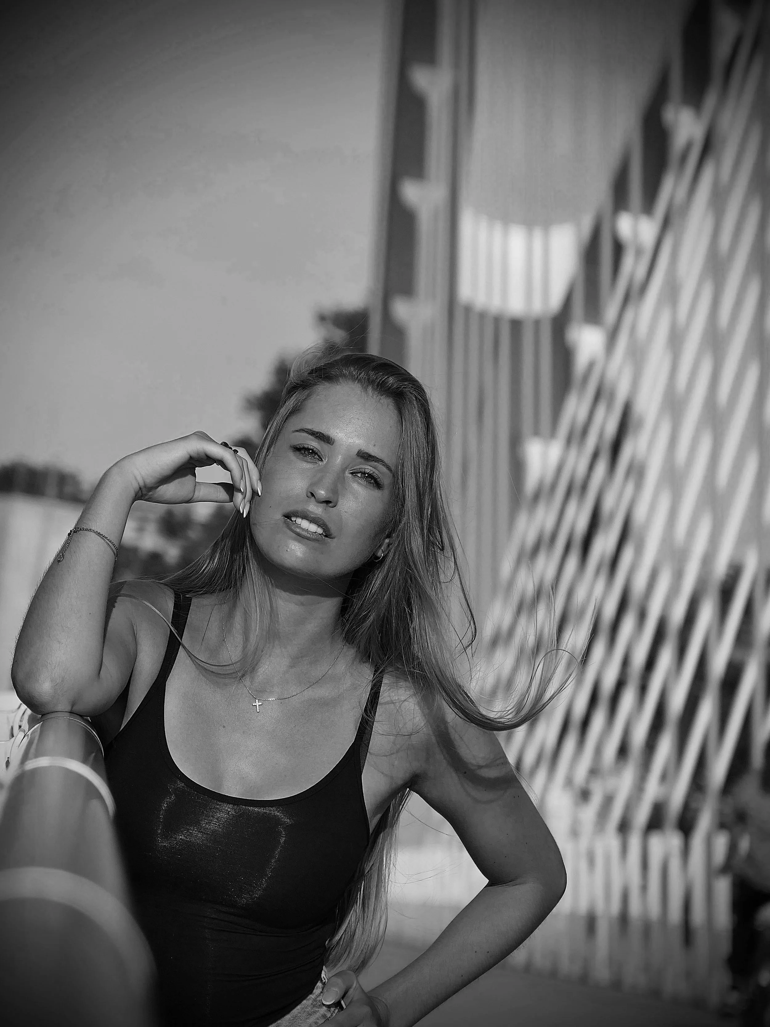 Jeune femme avec longue chevelure blonde, portant un débardeur noir, posant devant une structure moderne en métal, avec un ciel clair en arrière-plan.