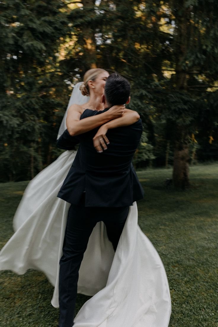 Un couple de mariés danse dans un parc verdoyant, l'homme portant un costume noir et la femme une robe de mariée blanche, sous un arbre.