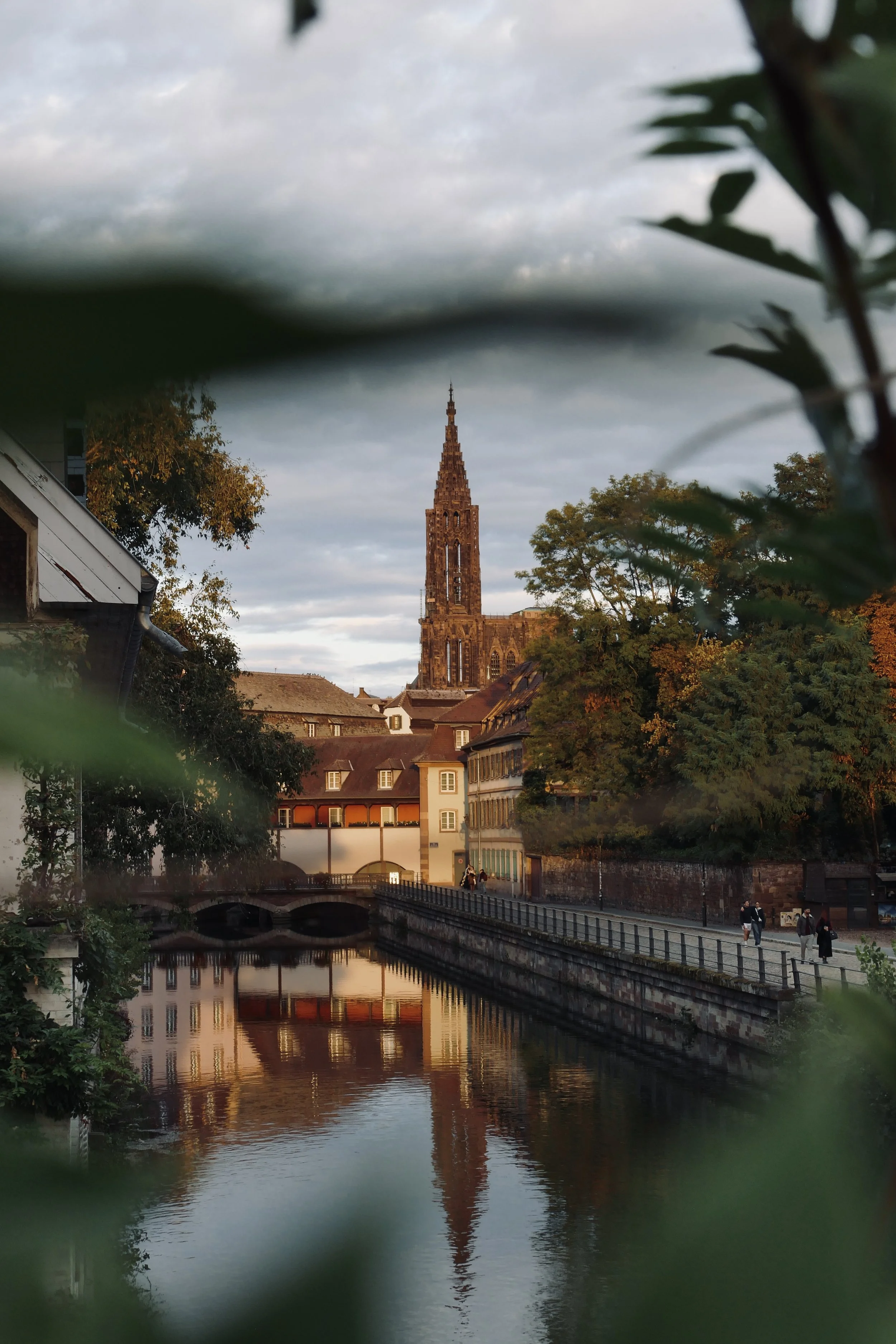 Vue d'une rivière avec un pont et des bâtiments, une église gothique en arrière-plan, arbres aux couleurs automnales, ciel nuageux, reflet dans l'eau.