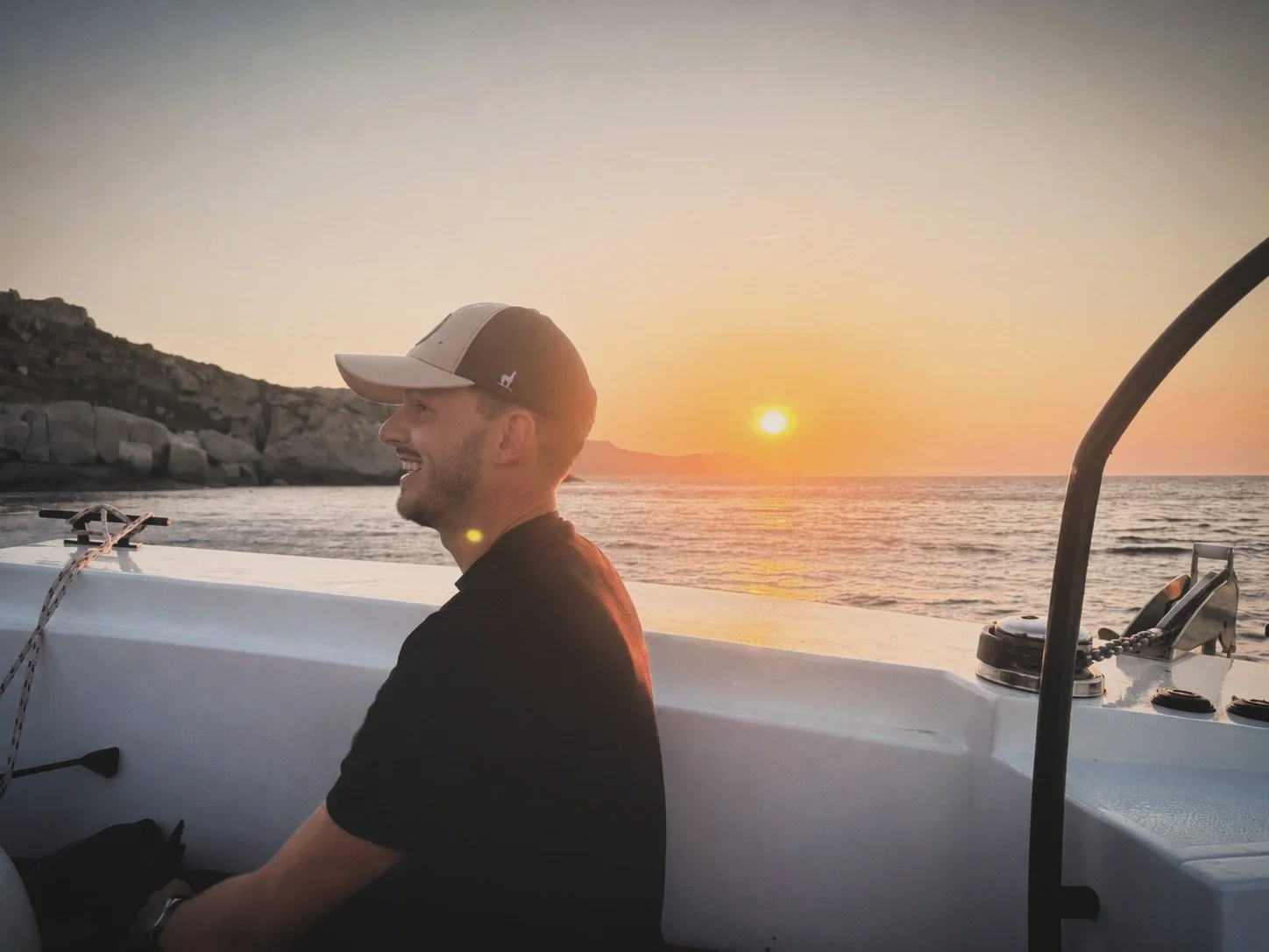Un homme souriant à capuche noire et casquette beige sur un bateau au coucher du soleil, avec la mer en arrière-plan