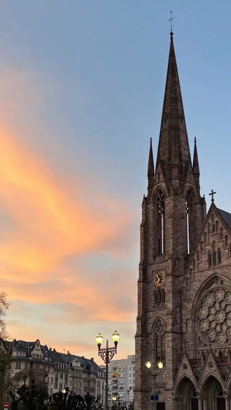 Une église en pierre avec une haute flèche, un cadran d'horloge, et des fenêtres gothiques, sous un ciel au coucher du soleil avec des couleurs orange et bleu.