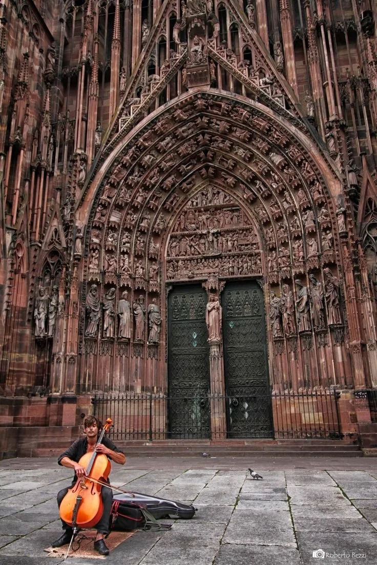 Un musicien jouant du violoncelle devant une grande porte en pierre ornée de sculptures religieuses, probablement une cathédrale gothique.