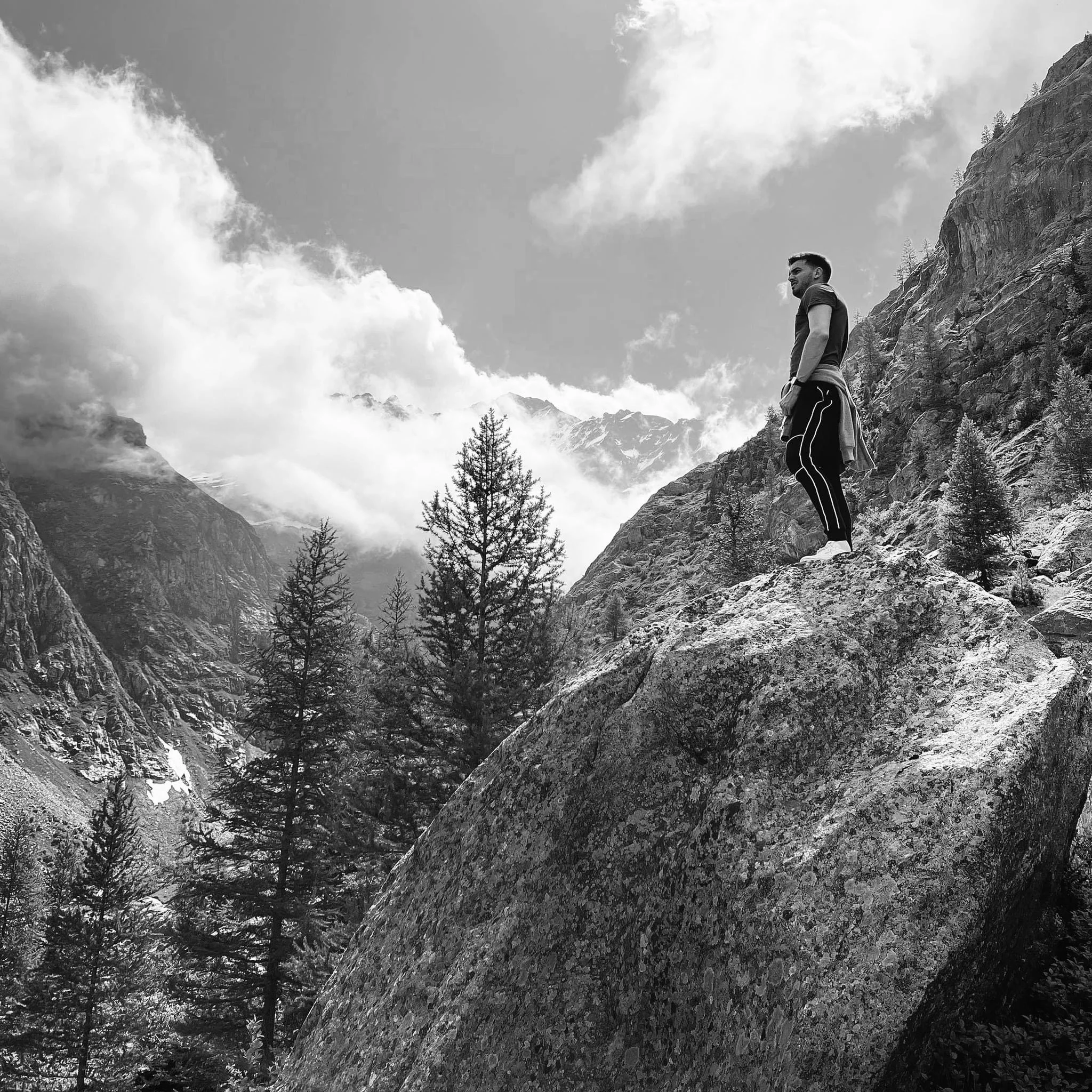 Homme debout sur un rocher dans une chaîne de montagnes avec arbres et nuages au ciel.