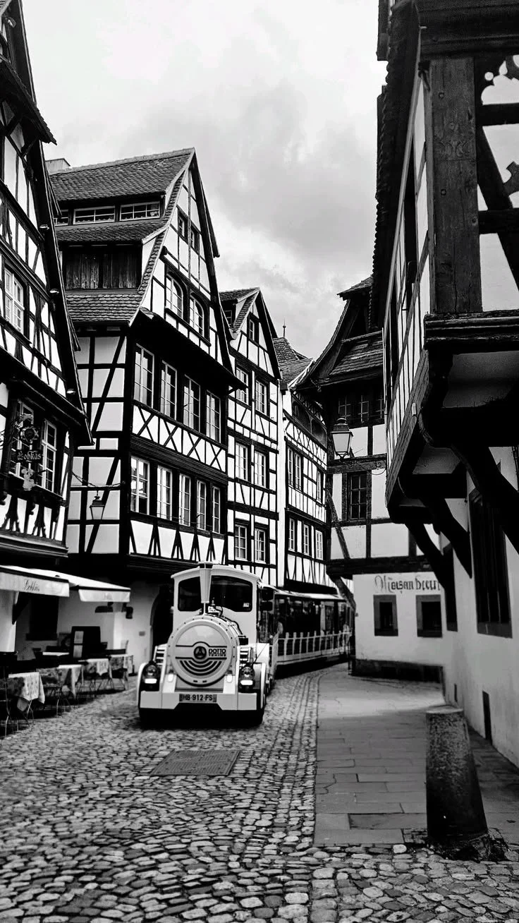 Vue d'une rue pavée avec des bâtiments à colombages typiques de l'architecture alsacienne, un petit train touristique et des lampadaires en fer forgé.
