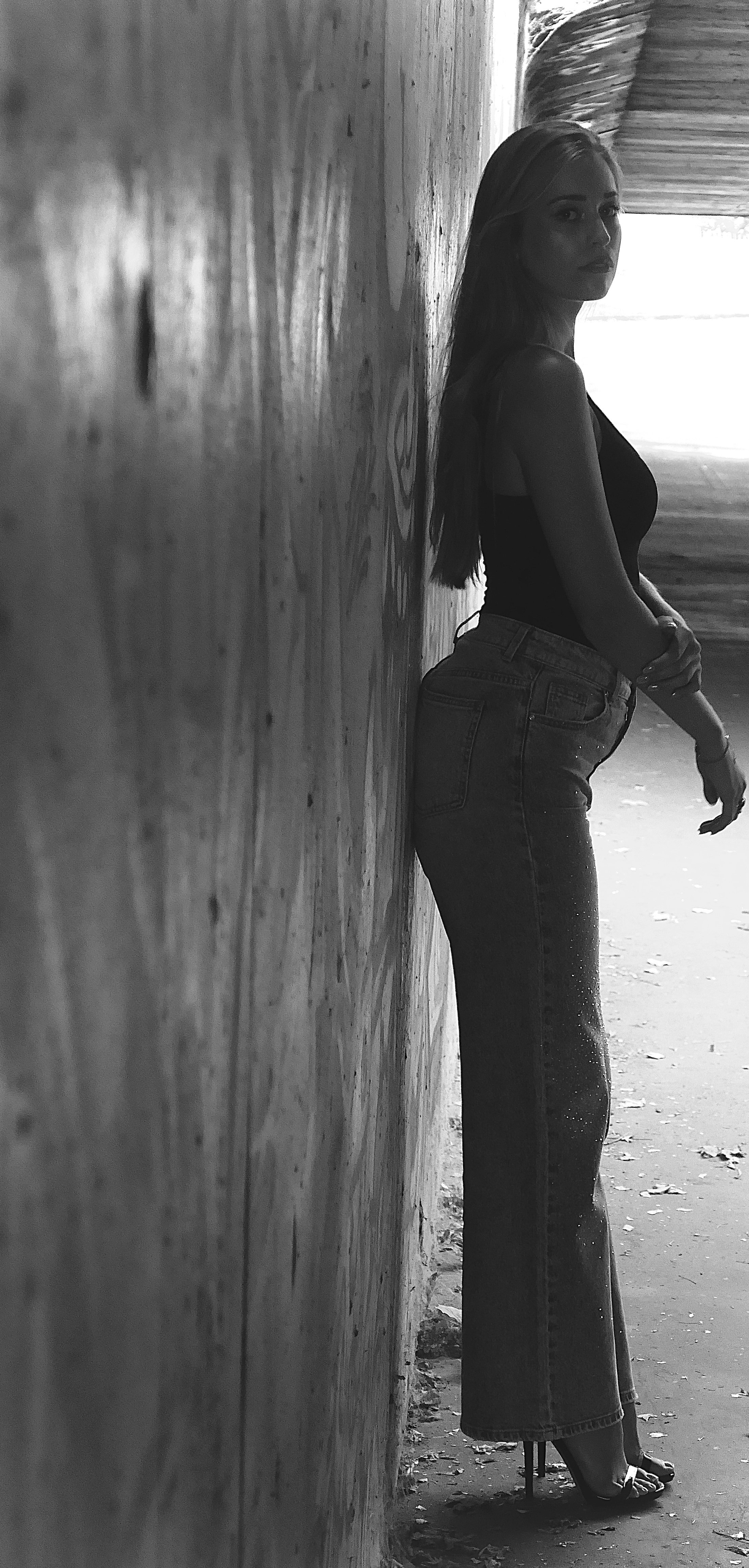 Femme debout contre un mur en bois, vue de profil, portant un t-shirt noir, un pantalon large taille haute et des sandales à talons, dans un espace lumineux avec un fond flou.