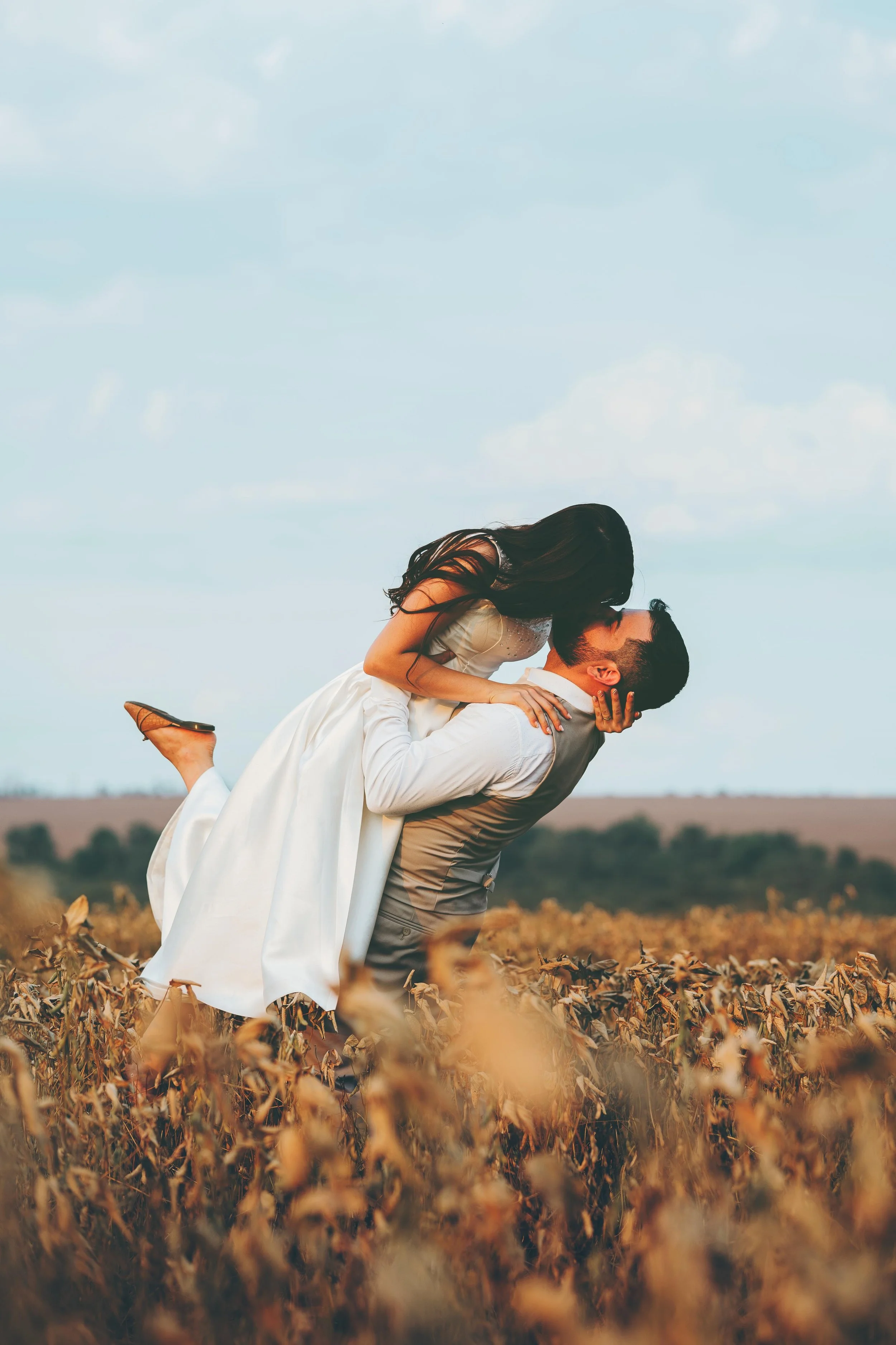 Un homme portant un costume donne une étreinte à une femme en robe de mariée dans un champ de maïs, sous un ciel bleu avec quelques nuages.