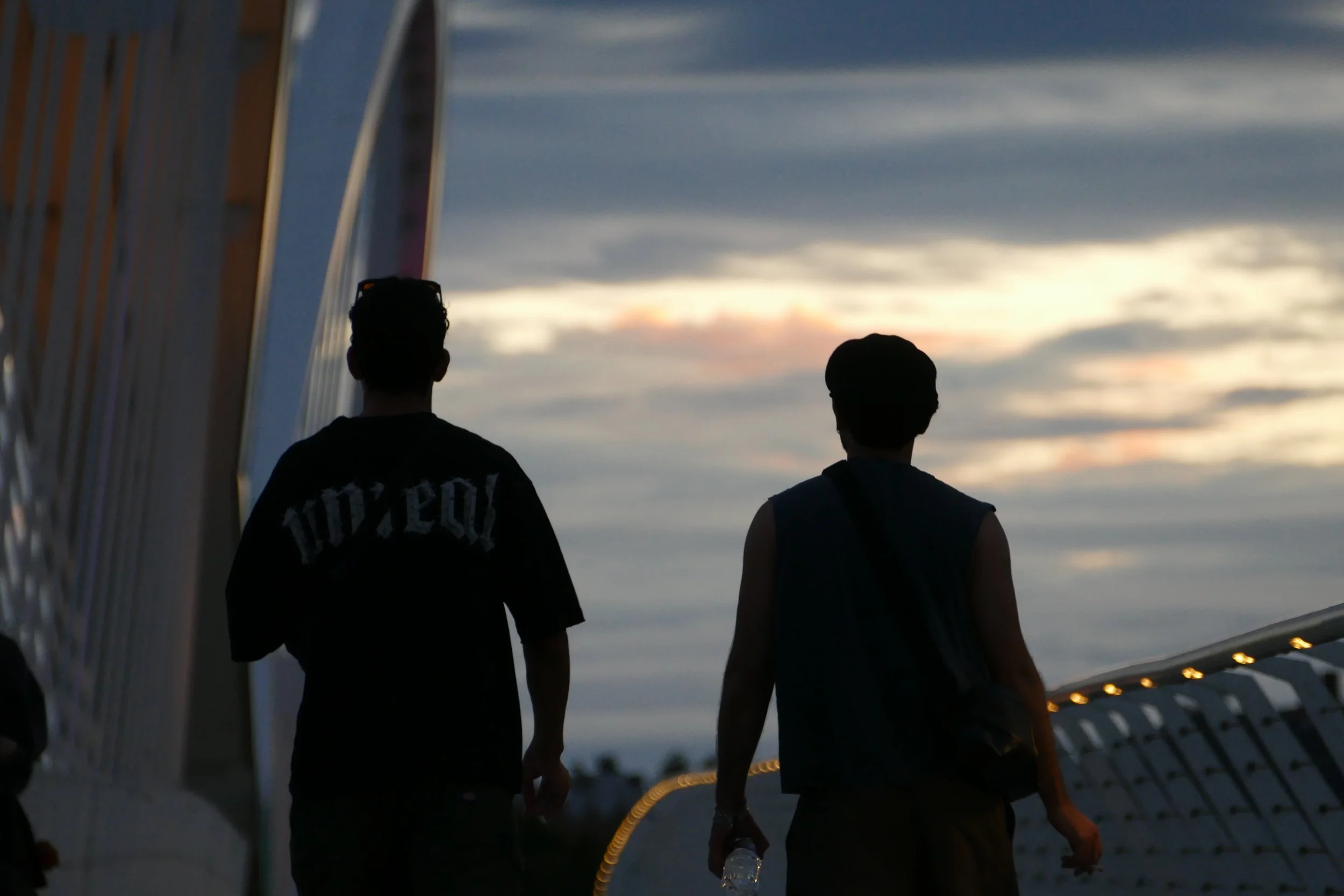 Photographie de deux silhouettes marchant sur une passerelle à Strasbourg au coucher du soleil, avec un ciel nuageux en arrière-plan.