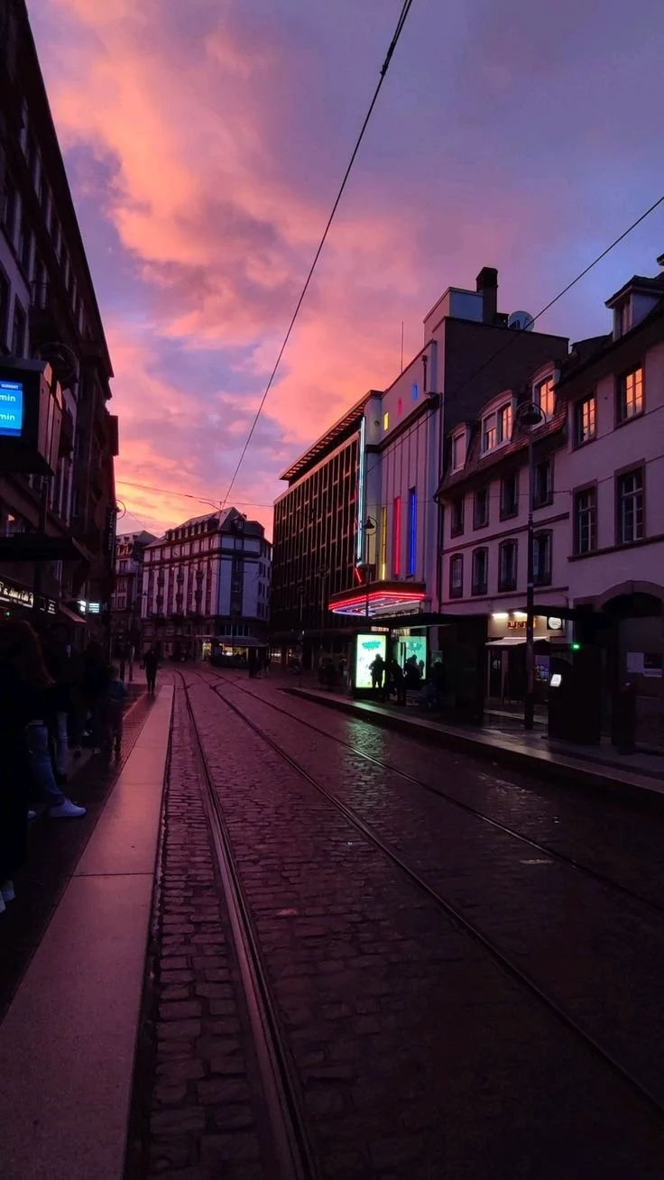 Une rue urbaine calme au crépuscule avec un ciel aux nuances rose et violet, des bâtiments modernes et anciens, des passants et des tramways électriques.