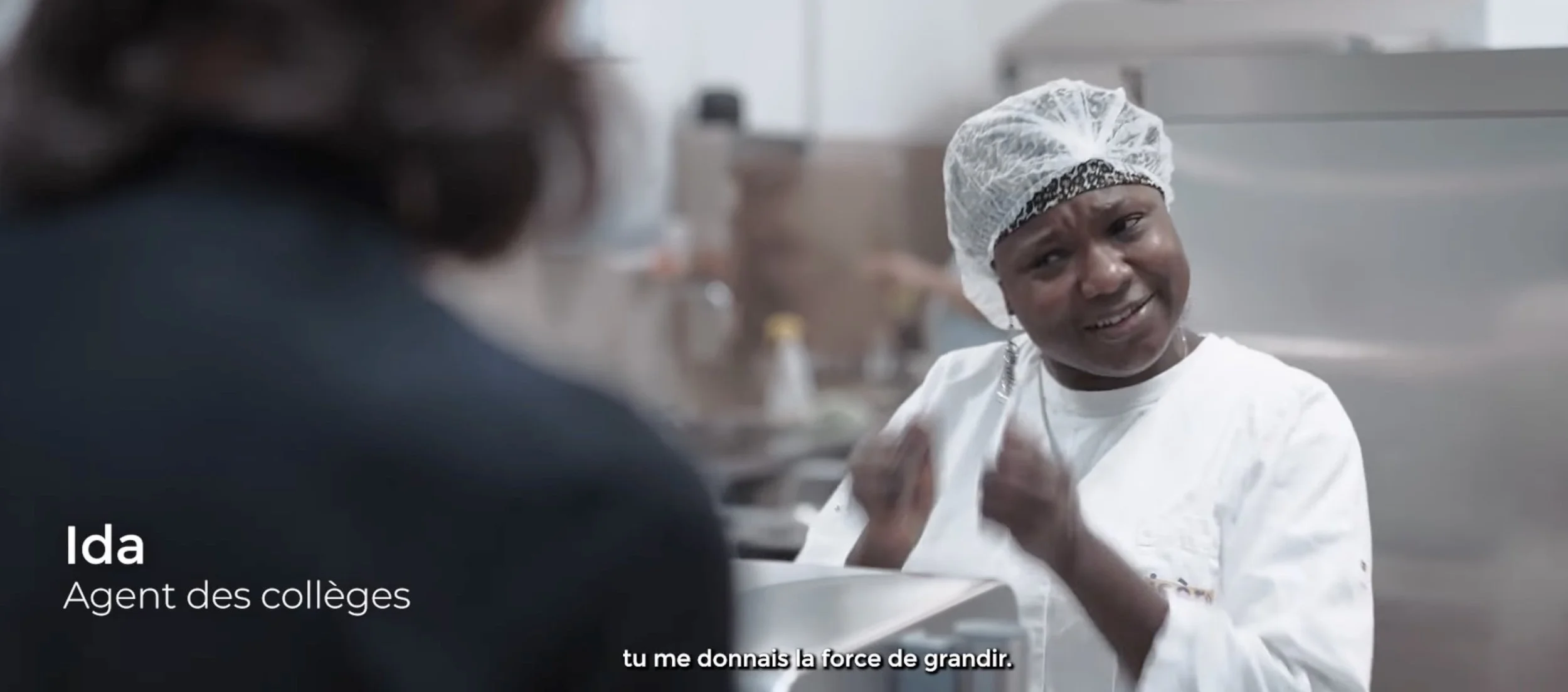 Une femme portant un uniforme de cuisine et un bonnet de chef parle à une autre personne. Elle sourit et semble encourager. Texte superposé : "Ida Agent des collèges" et sous-titre : "tu me donnes la force de grandir".