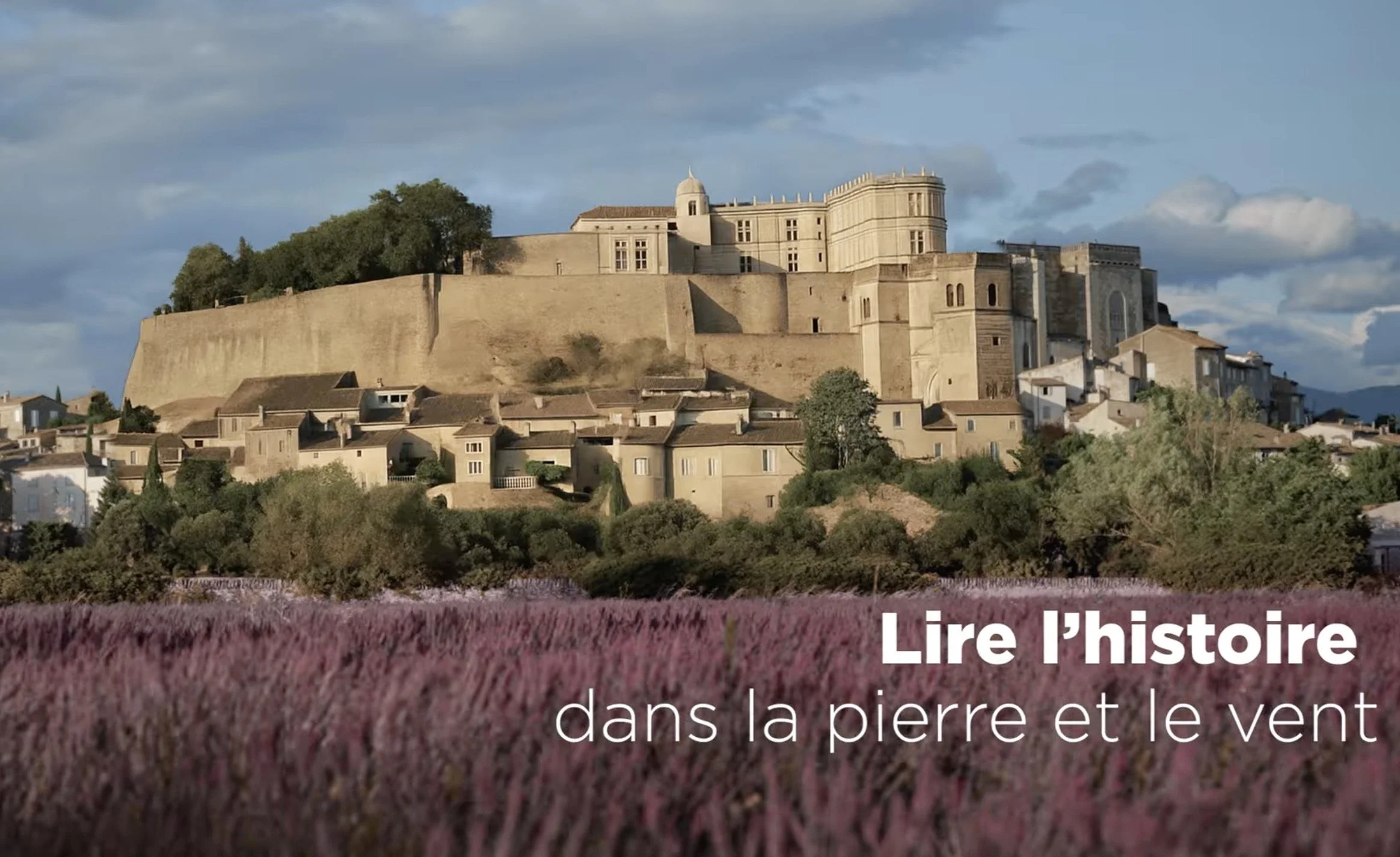 Un château perché en haut d'une colline entourée d'arbres, avec un ciel partiellement nuageux, et une forêt de lavande en premier plan.