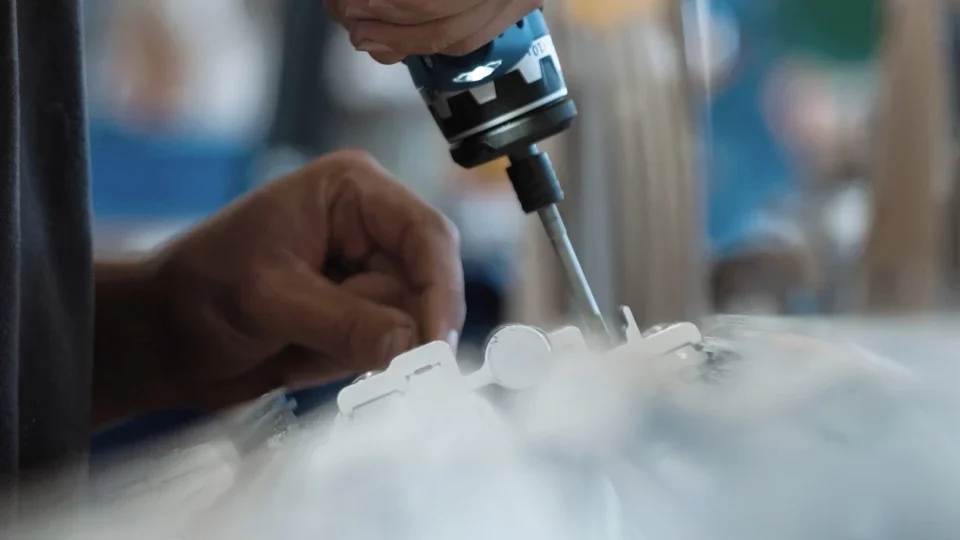 Une main manipule un tournevis pour fixer une pièce en plastique blanche, une perceuse électrique est également visible.
