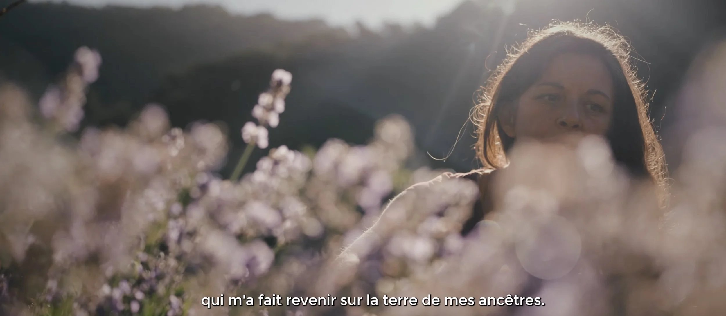 Une jeune femme dans un champ de fleurs, regarde vers l'objectif, éclairée par la lumière du soleil en arrière-plan.