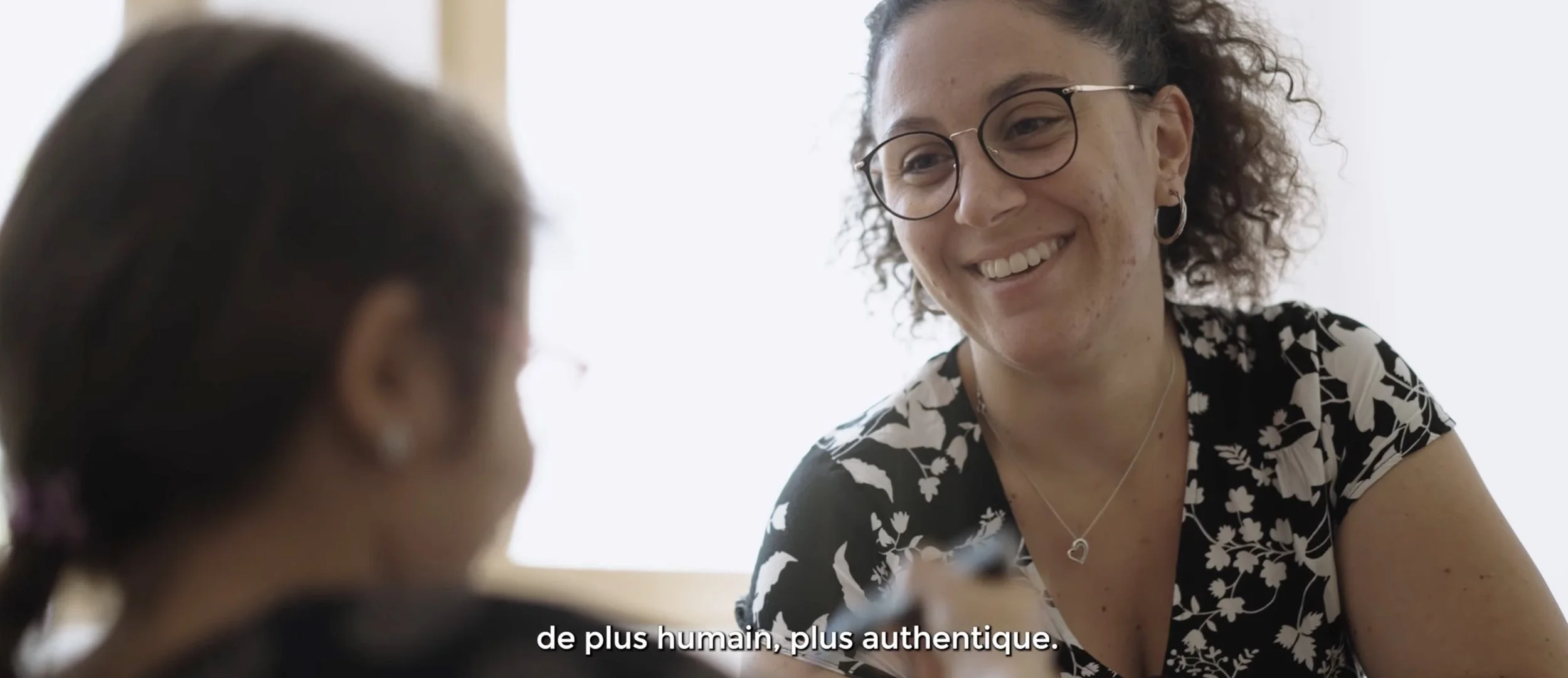 Une femme souriante porte des lunettes et parle à une personne dont la tête est visible au premier plan, dans un environnement lumineux avec des sous-titres en français.