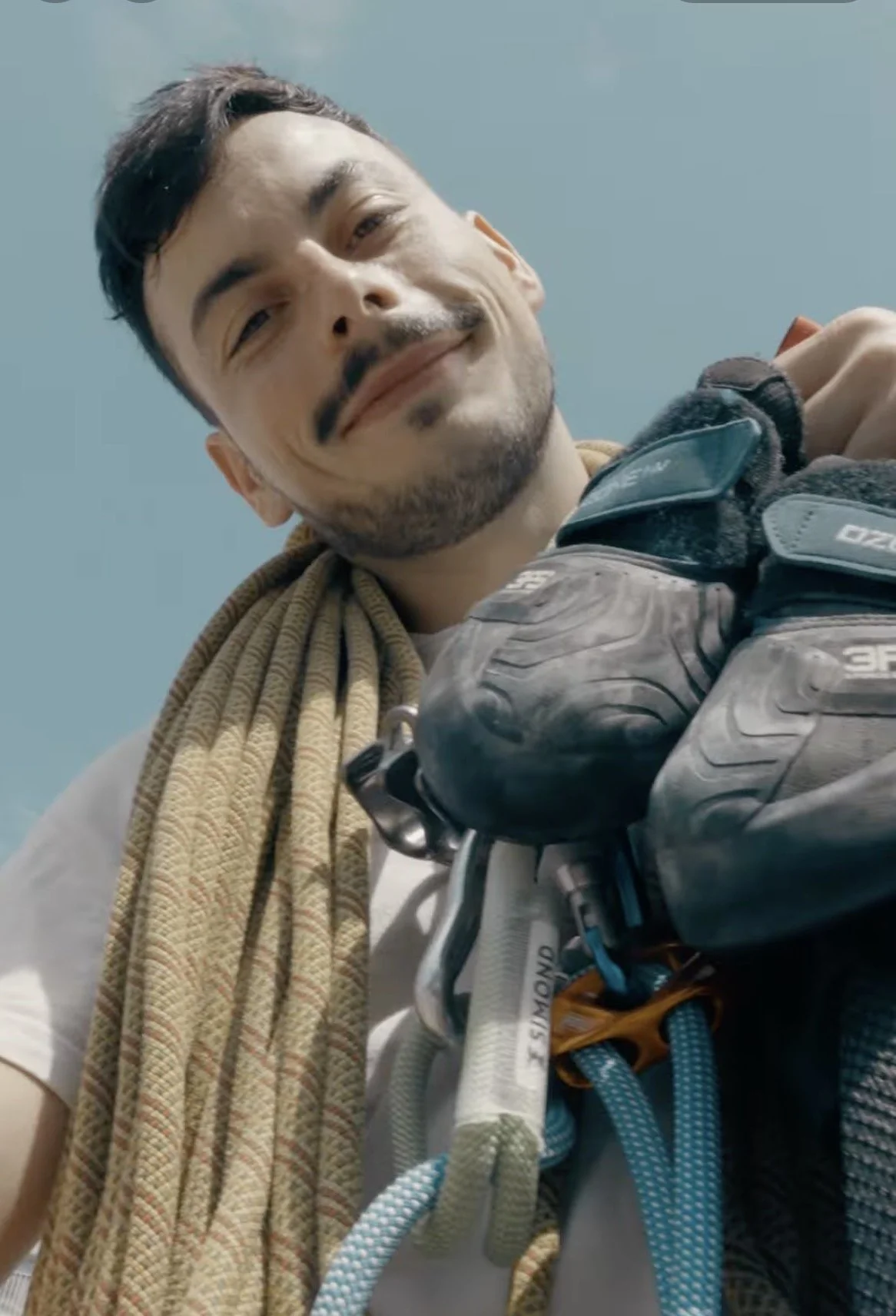 Homme souriant portant un casque d'alpinisme en montagne, avec un ciel bleu en arrière-plan.