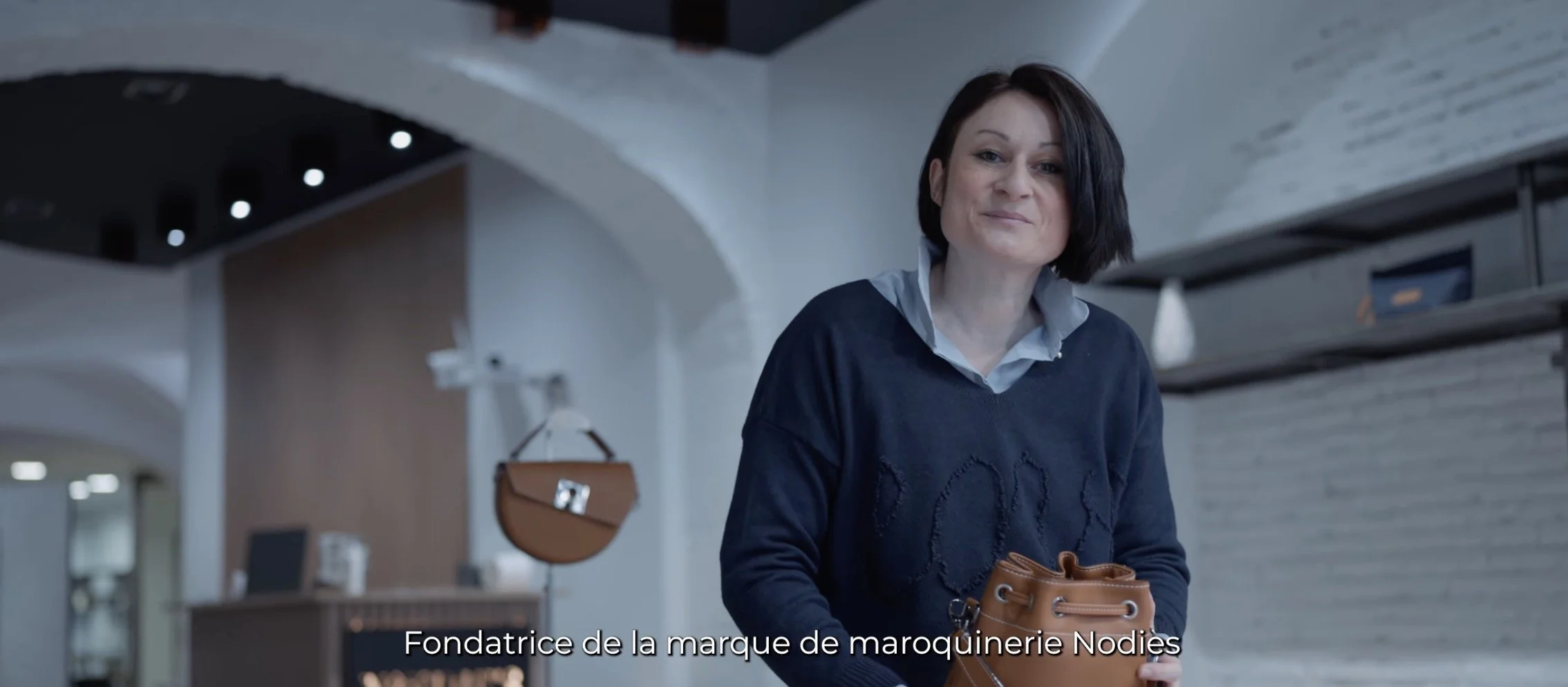Une femme avec des cheveux courts noirs, portant un pull bleu foncé et une chemise blanche, tenant un sac en cuir brun dans un intérieur moderne. Subtitles en français : "Fondatrice de la marque de maroquinerie Nodies".