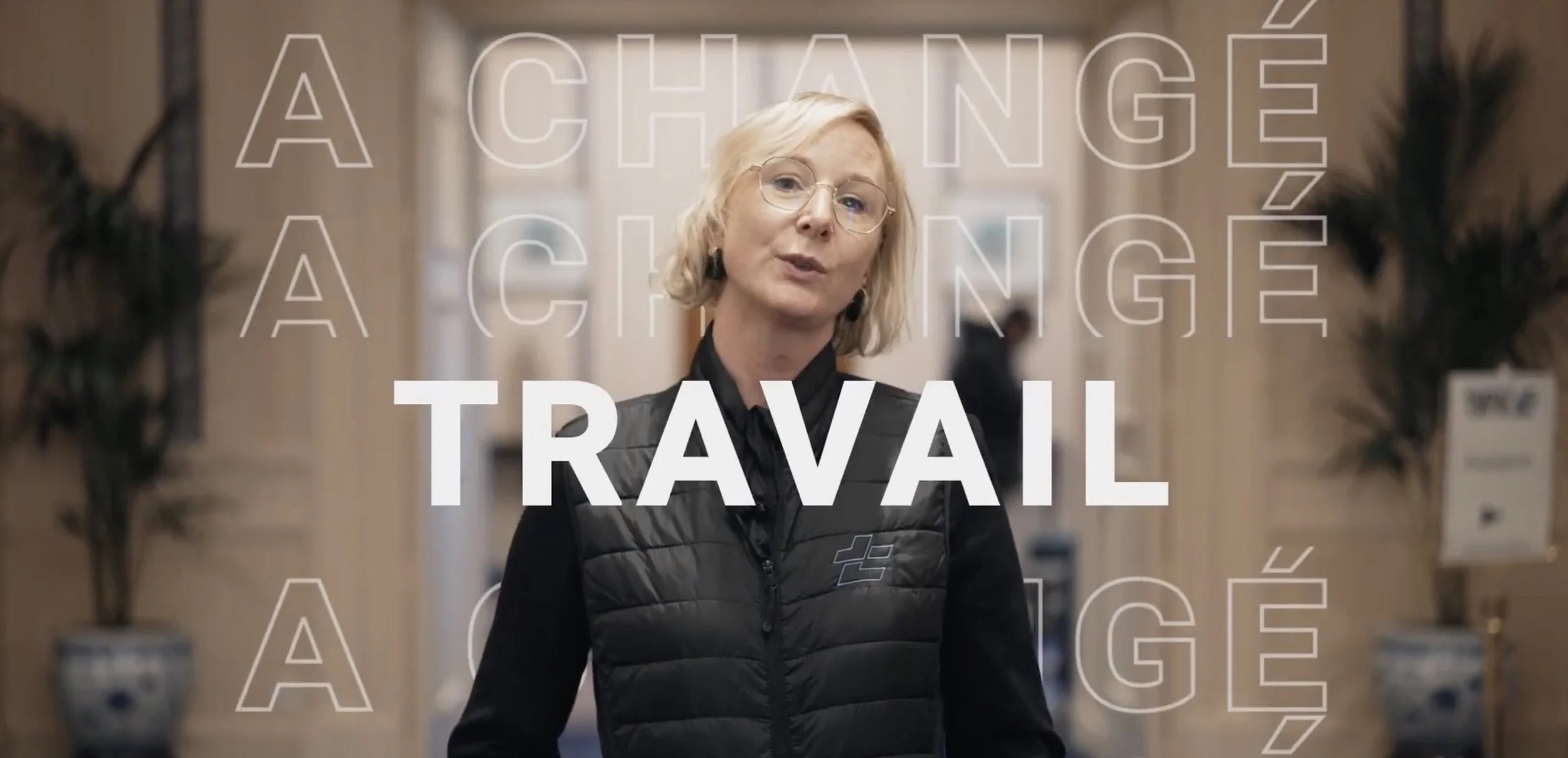Une femme blonde portant des lunettes et un gilet noir se tient dans un endroit intérieur, probablement une boutique ou un bureau, avec du texte en français sur l'écran disant 'À CHANGER, À CHANGER, TRAVAIL'.