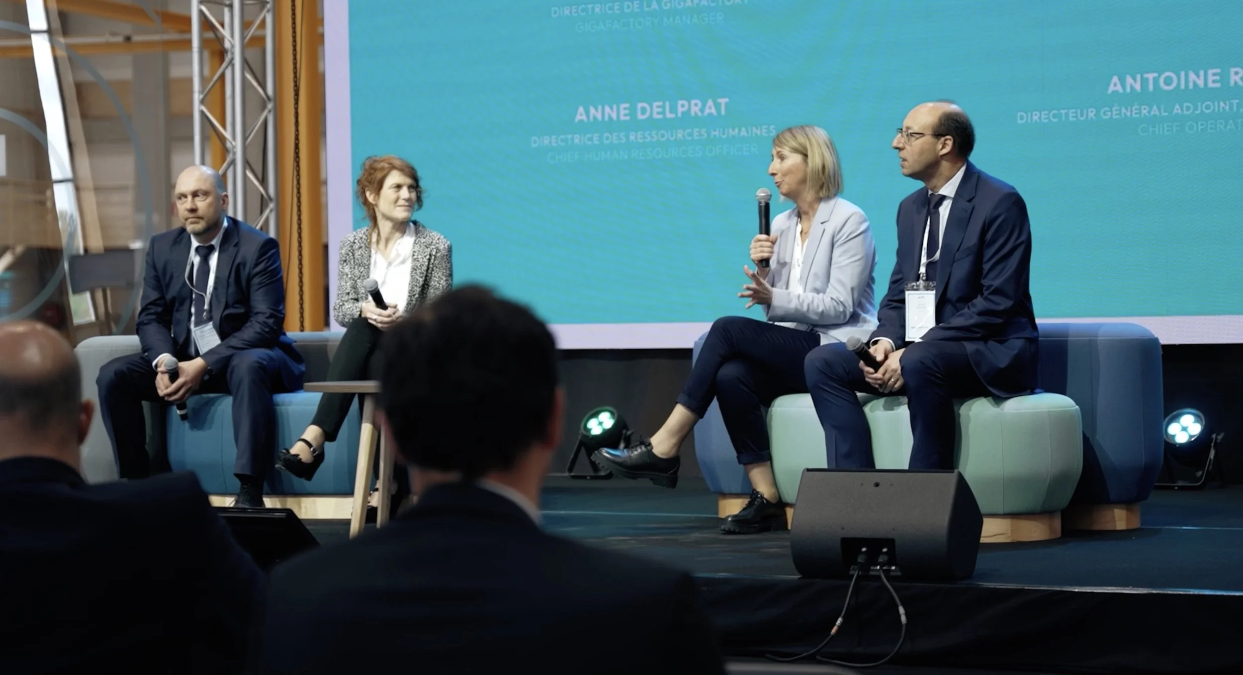 Quatre personnes assises sur un canapé lors d'un panel de discussion à une conférence, avec un écran bleu en arrière-plan affichant des noms et titres en français et anglais.