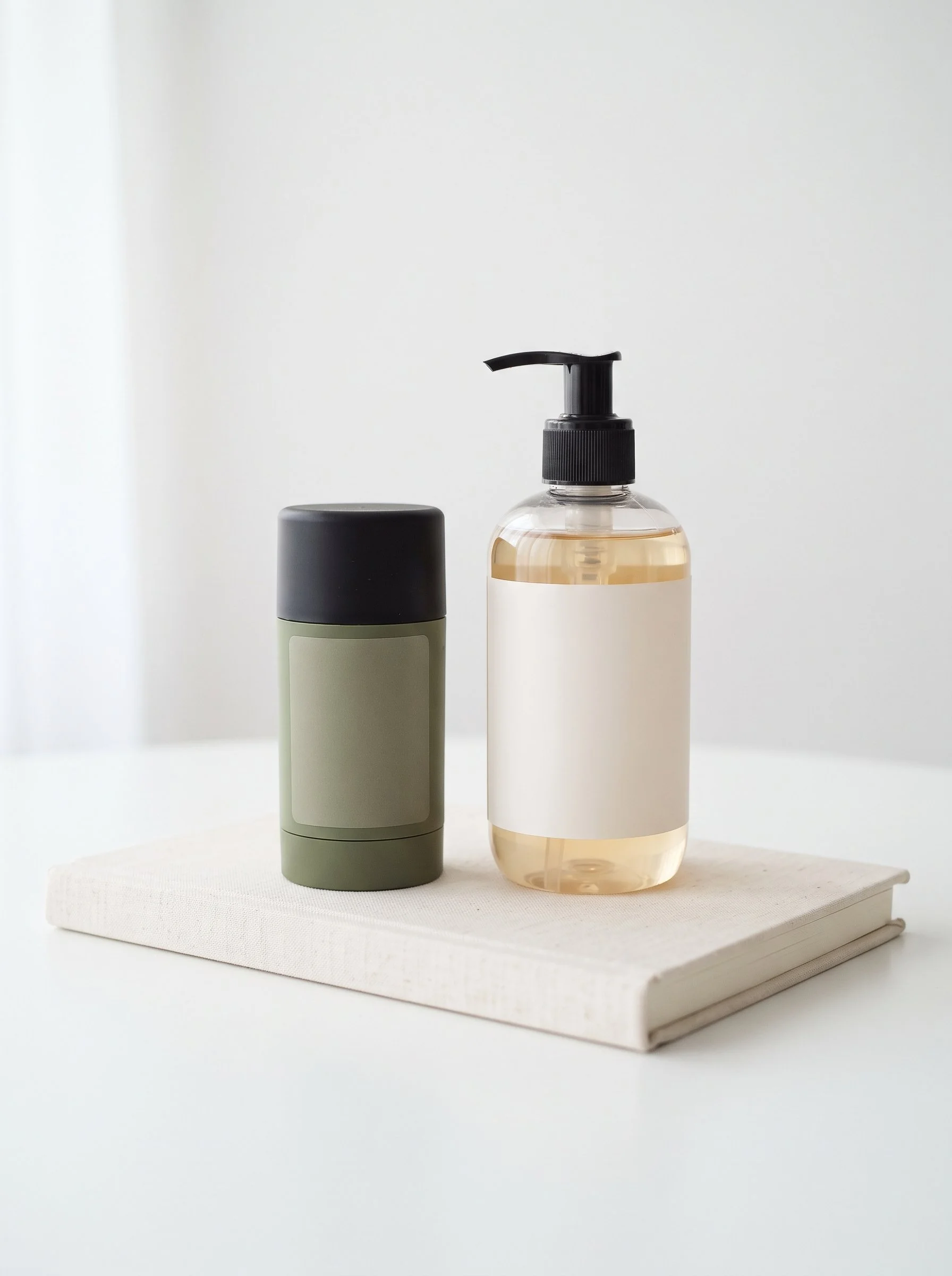 Two bottles, one with a pump dispenser and the other with a screw cap, placed on a white surface with a white background. The bottle with the pump dispenser is transparent and contains a light-colored liquid. The bottle with the screw cap is opaque gray and pale green.