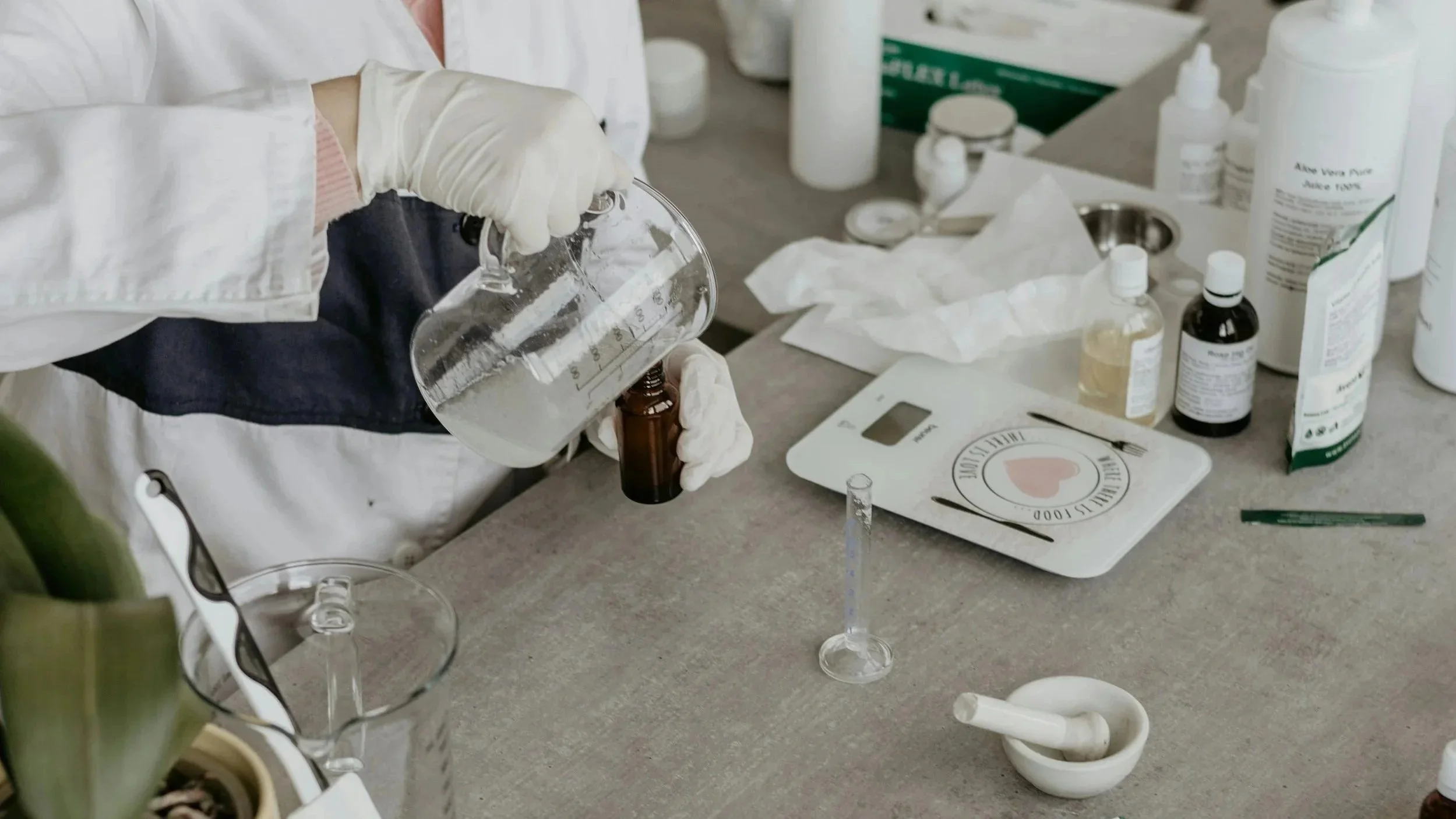 Person wearing white gloves pouring liquid into a small dark vial in a laboratory setting with various bottles, syringes, and measurement tools on the table.