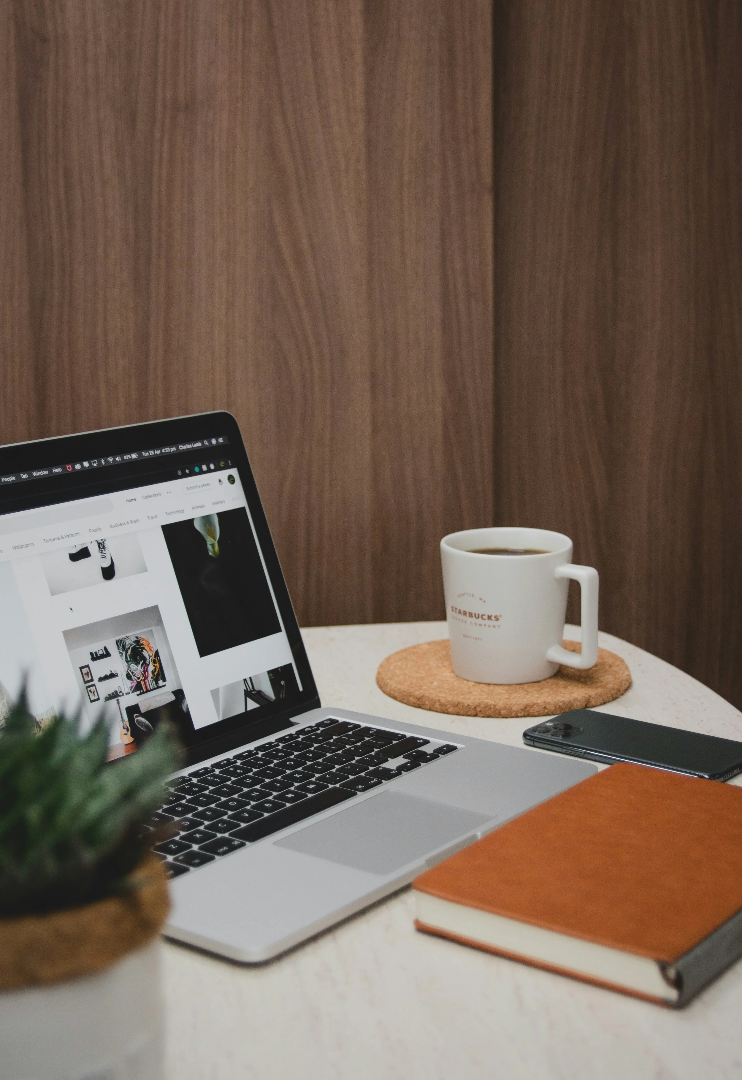 A close-up of a workspace with a silver MacBook showing a web page, a white coffee mug with Starbucks logo, a brown leather notebook, an orange notebook, a smartphone, and a blurred plant in the foreground. The background features a wooden wall.