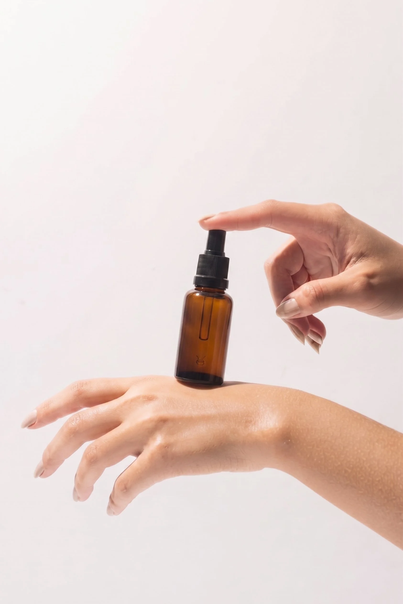 A person's hand applying a spray from a small amber glass bottle onto the back of another person's hand against a plain white background.