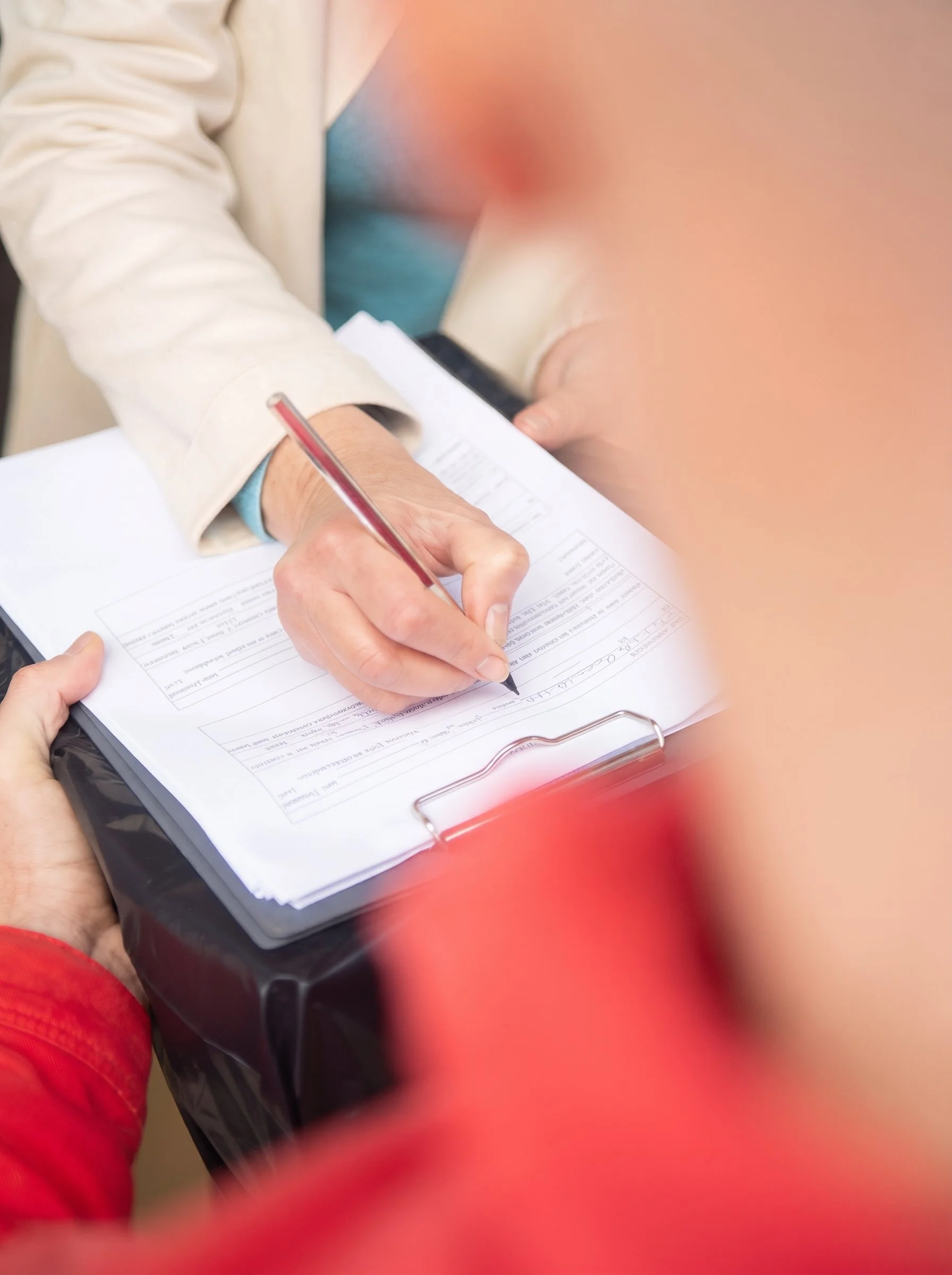 Person signing a document with a pen on a clipboard, with other hands holding the clipboard steady. The person is wearing a red shirt and beige jacket, and the setting appears to be indoors.
