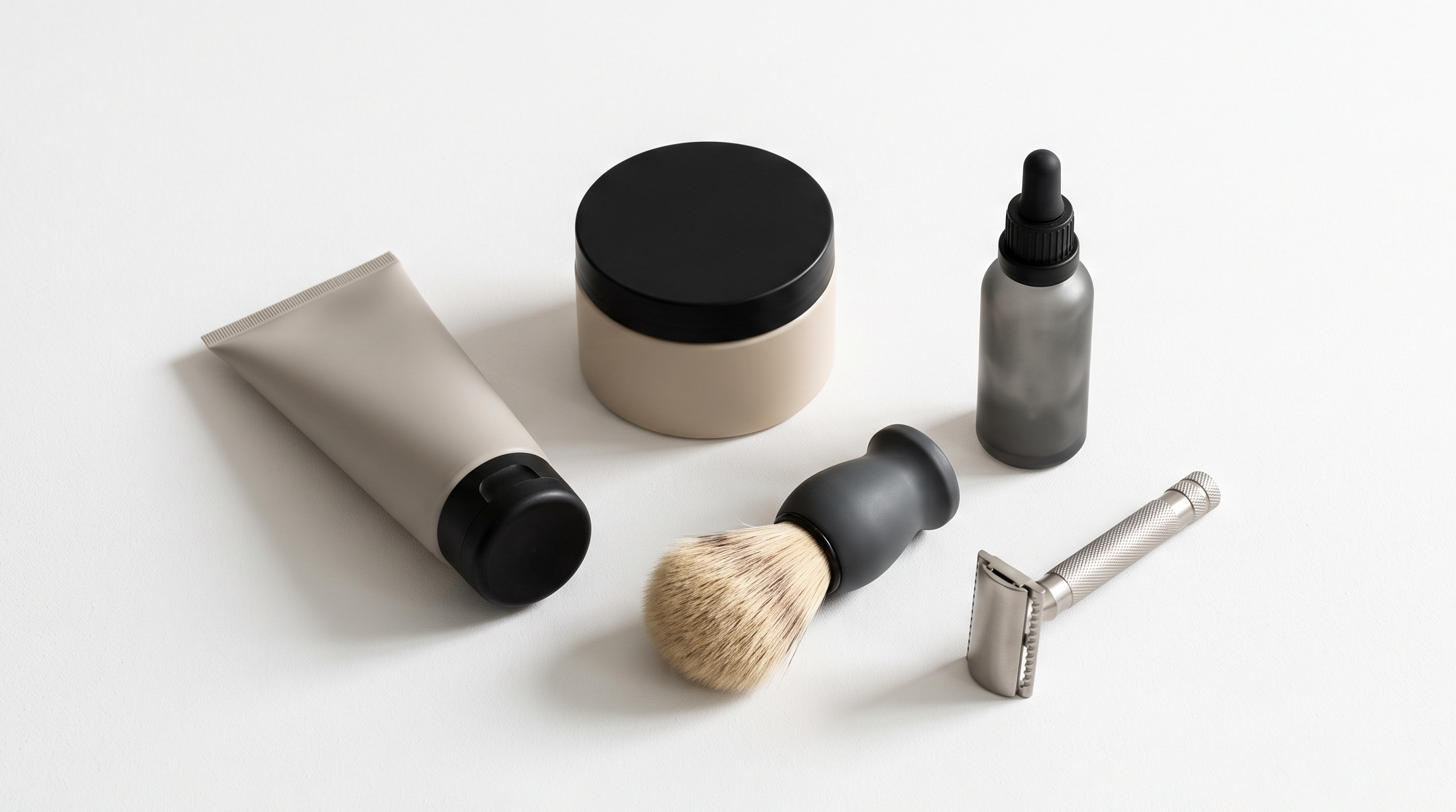 Collection of men's grooming products and shaving accessories on a white surface, including a tube of cream, a jar of balm, dropper bottle.