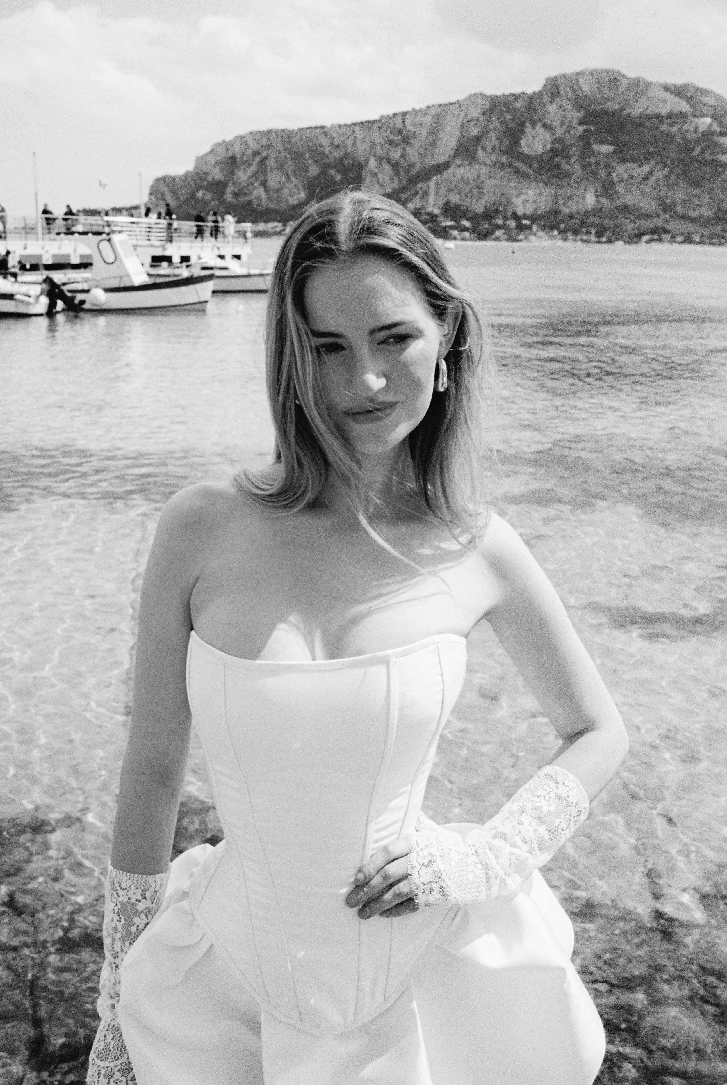 A woman in a white dress with lace sleeves poses on a beach with boats and a mountain in the background. Italian wedding, destination wedding
