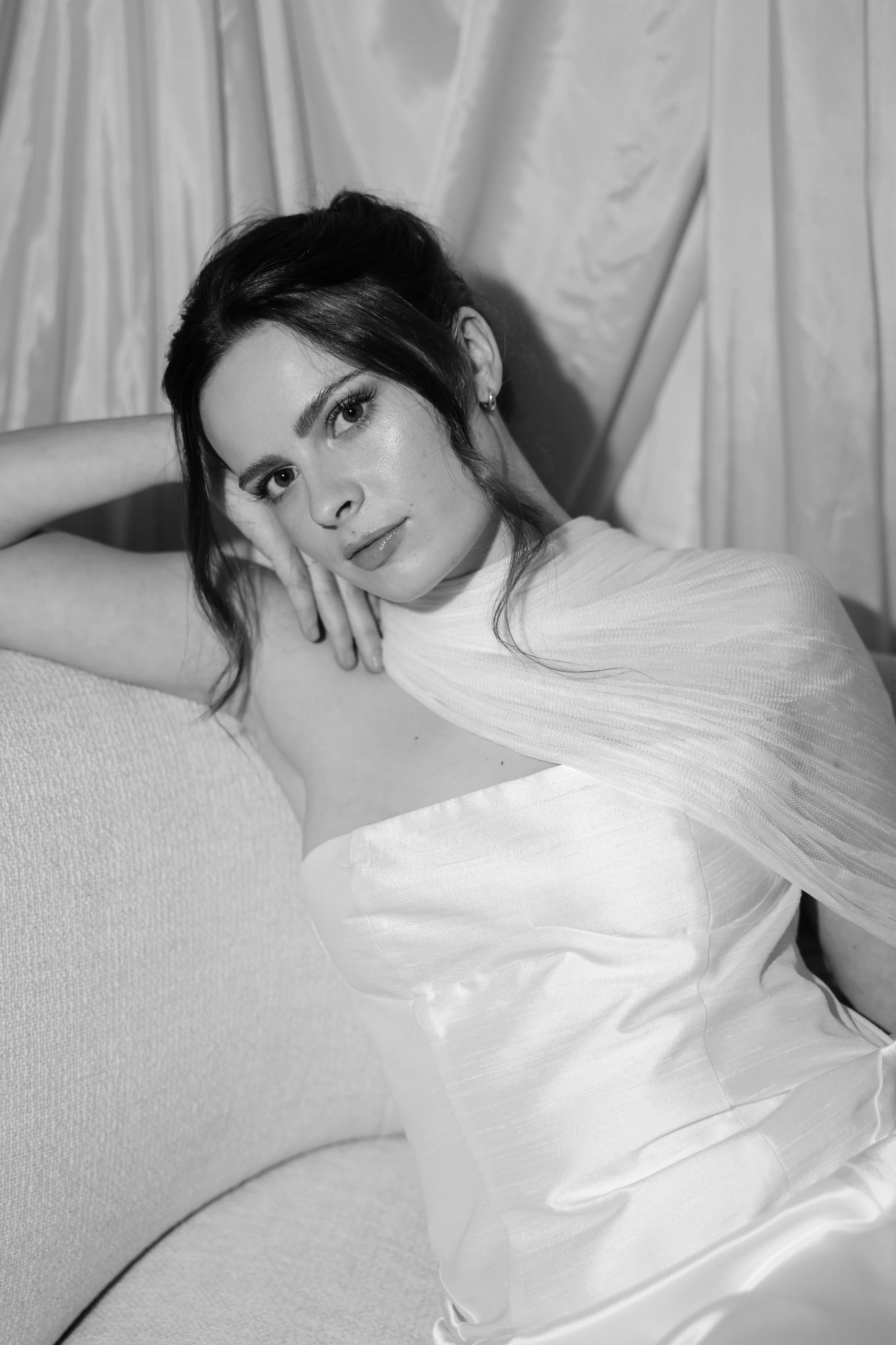 A woman with dark hair and earrings reclining on a surface with draped curtains in the background, wearing a light-colored dress, in a black-and-white photograph.