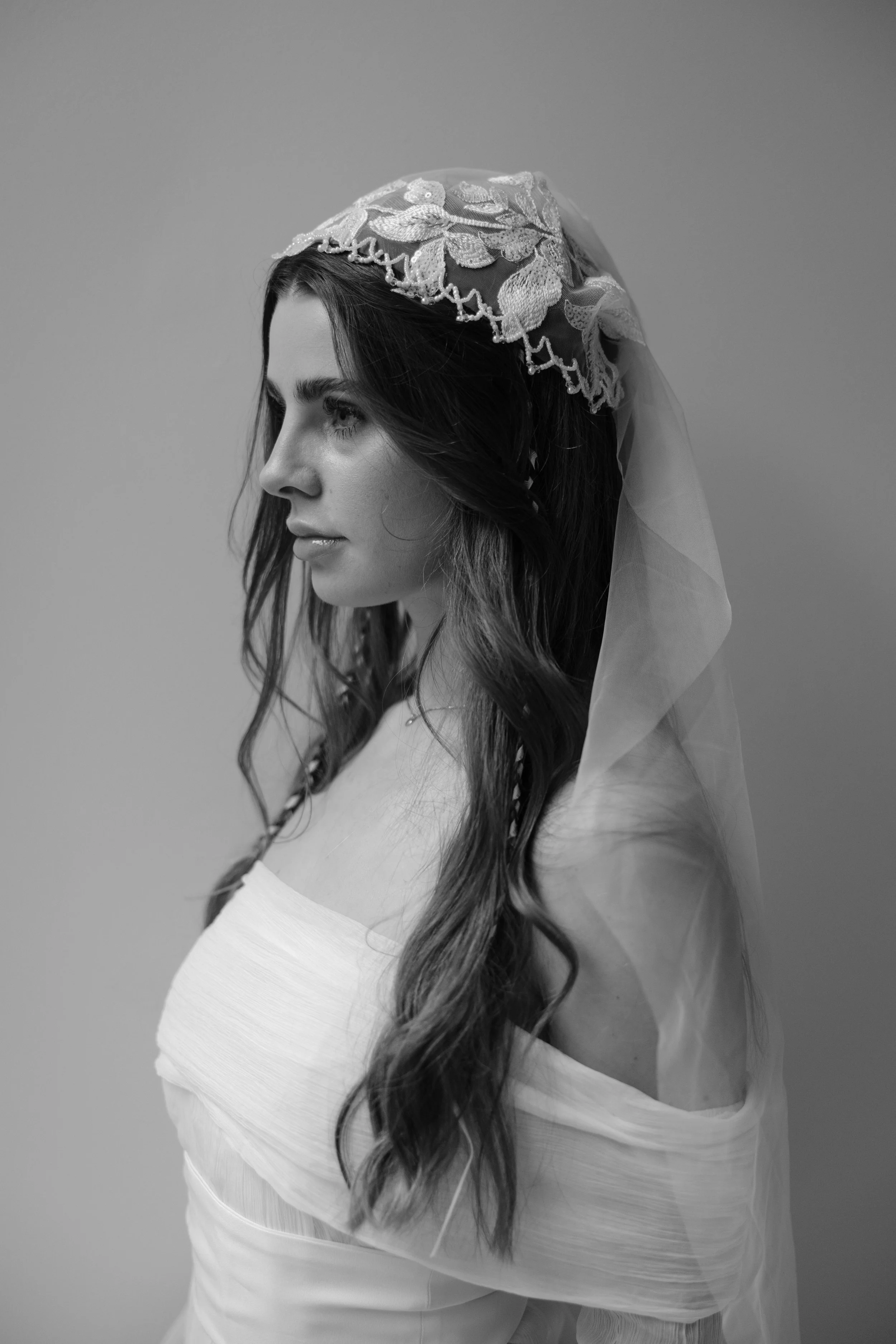 A black and white photograph of a woman with long wavy hair wearing a lace mantilla on her head, looking to the left against a plain background. Bride bridal beauty
