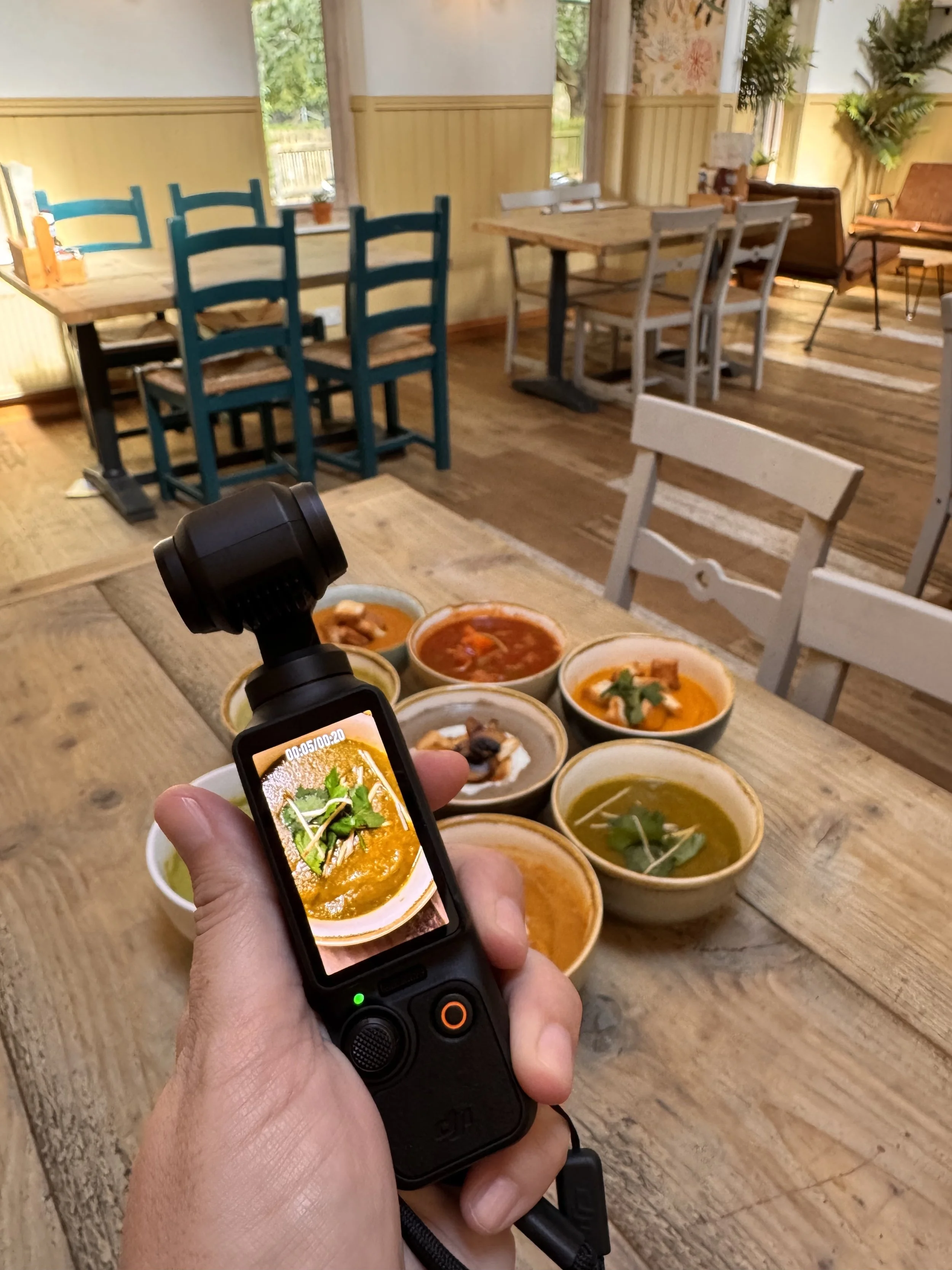 A hand holding a Dji Osmo Pocket 3 in the foreground, showing a bowl of soup on the screen, with several bowls of soup in the background